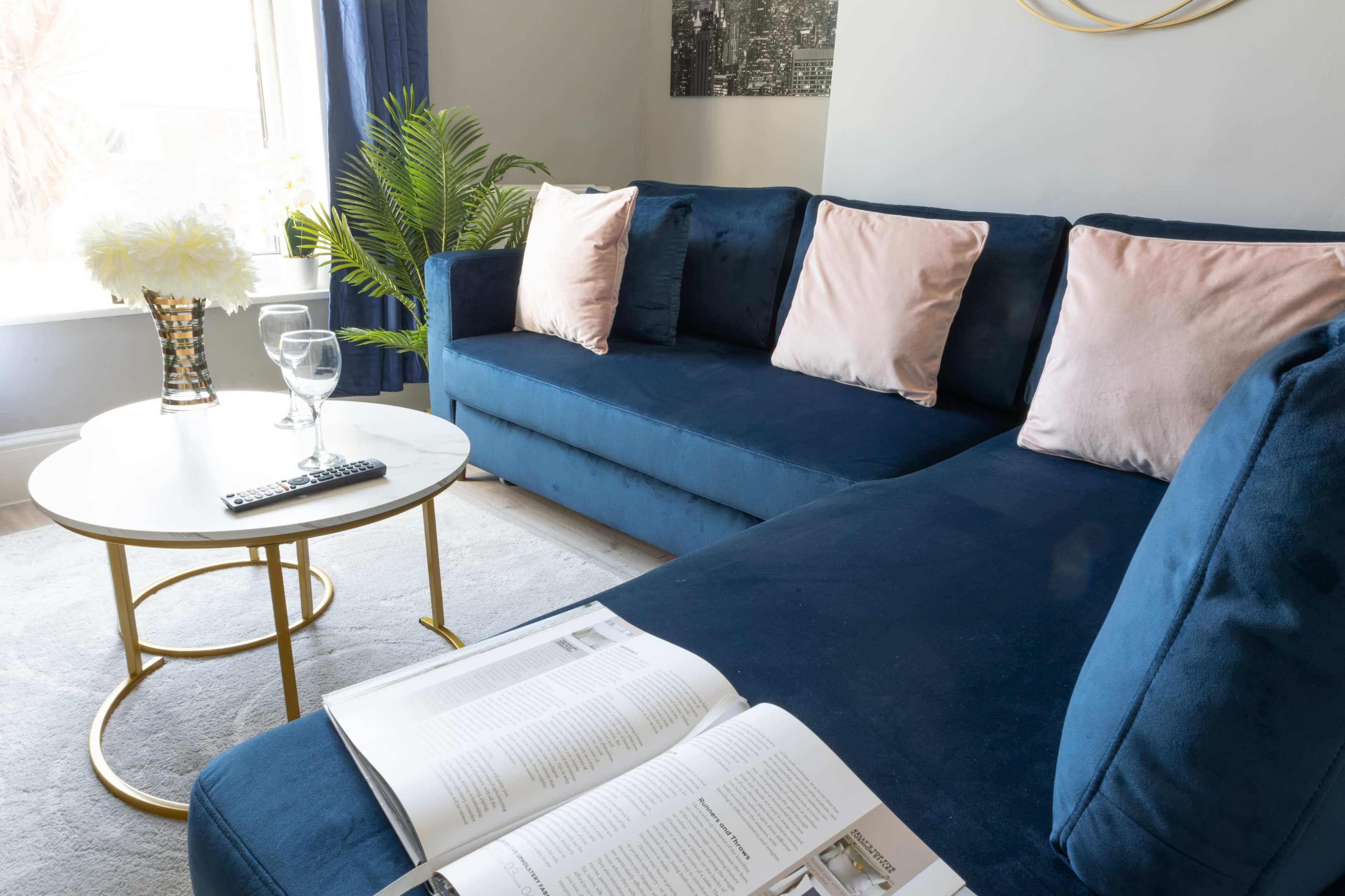 A blue velvet sectional sofa with pink cushions is positioned next to a round coffee table set with a glass and an open magazine.