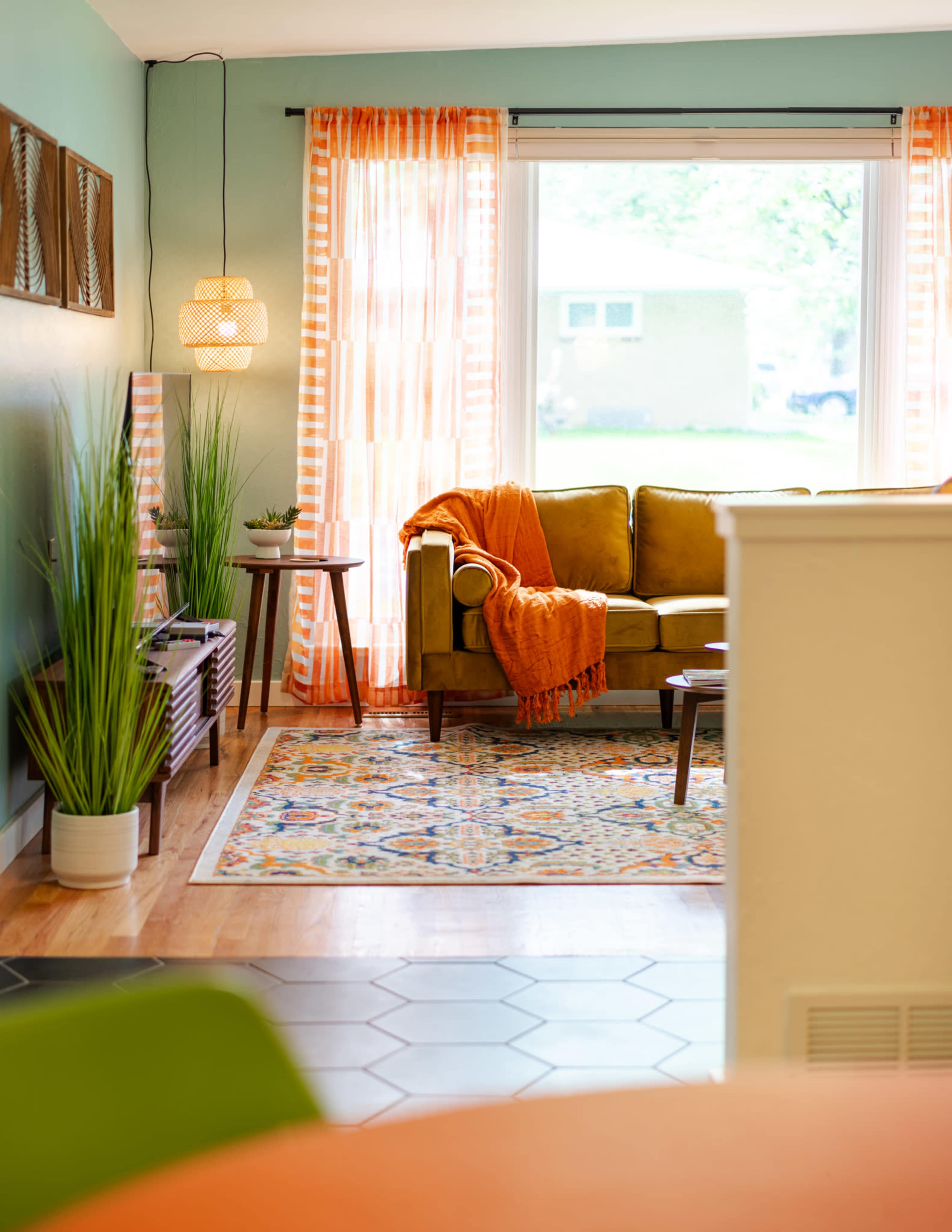 A bright living room features a green couch with an orange throw blanket, surrounded by plants and a patterned rug.