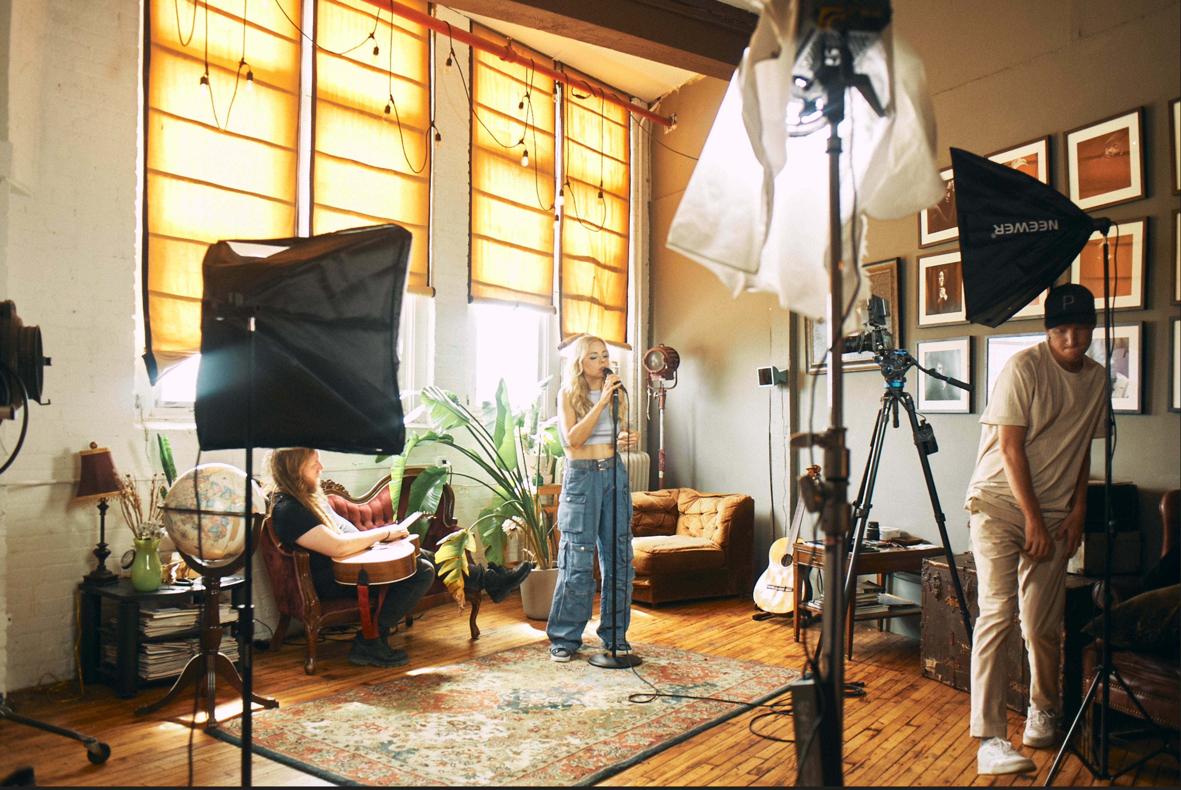 A woman stands in a well-lit room speaking into a microphone while another person sits nearby with a guitar and a third adjusts a camera setup.