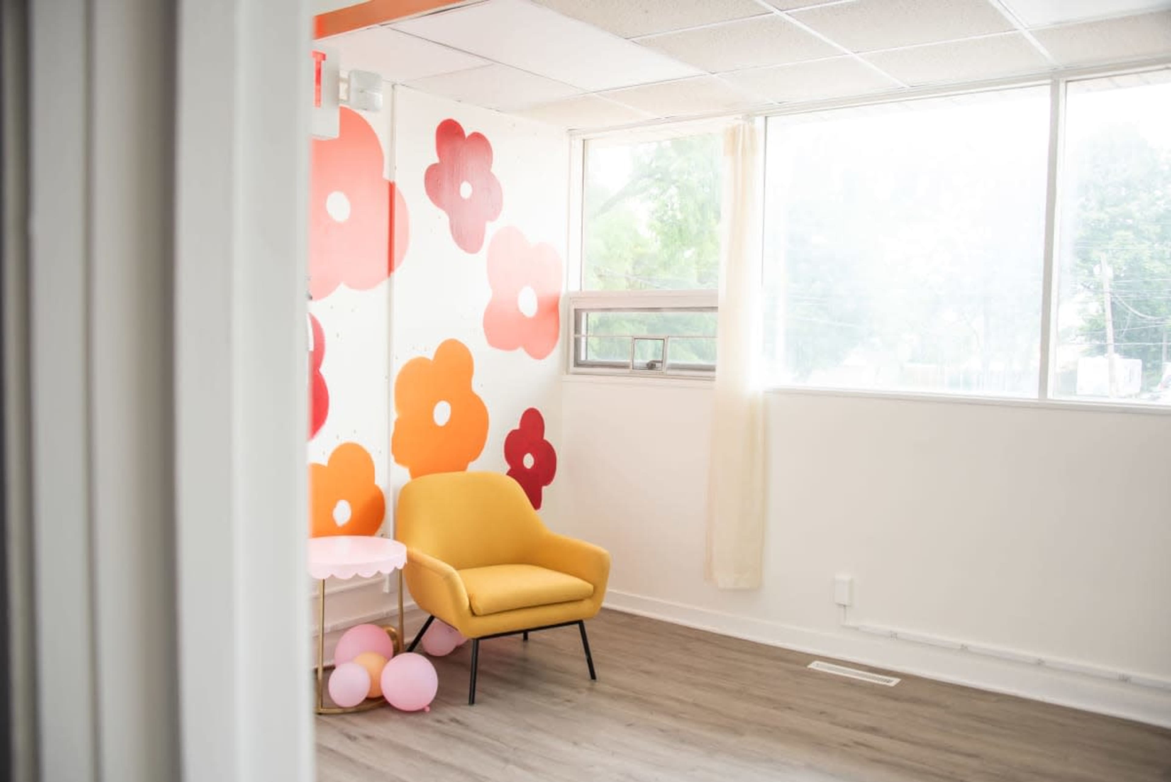 The image shows a bright, aesthetically designed room with a yellow chair, a small pink table, and a wall decorated with large red and orange flowers.