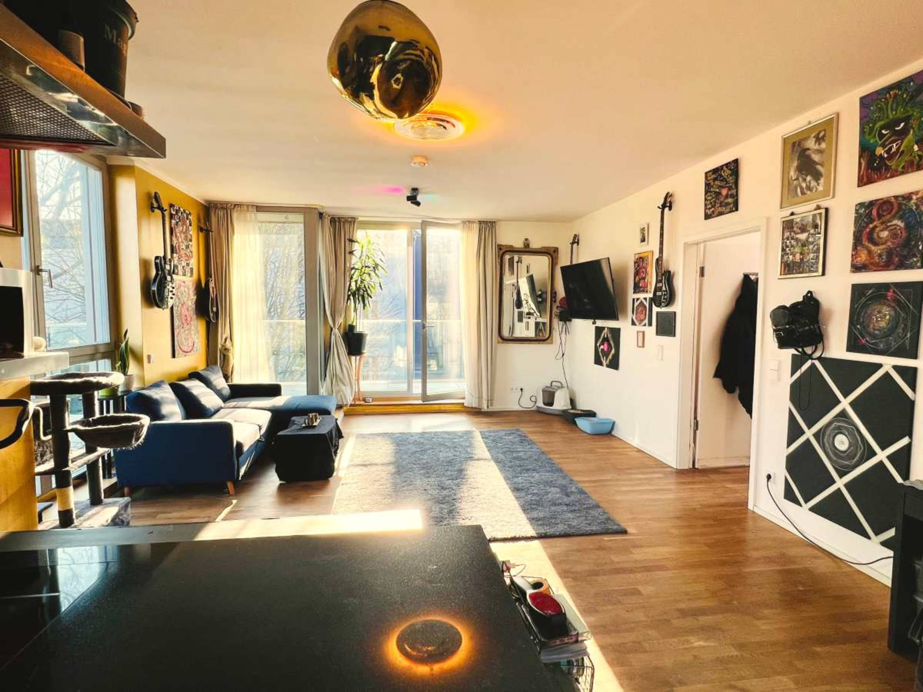 The image shows a modern, open living room with a blue sofa, large windows, and various artworks on the walls.