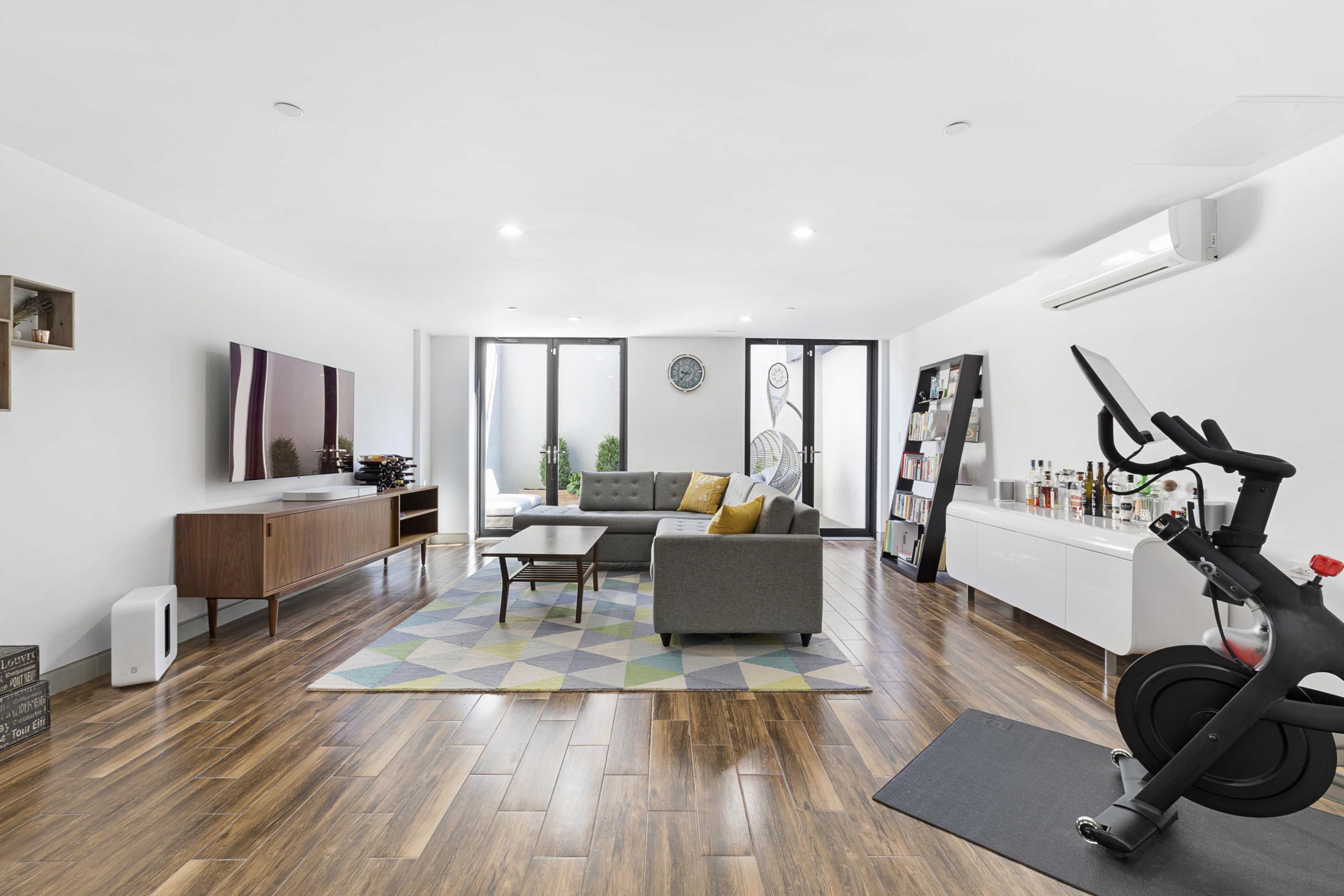 A modern living room with a grey sofa, wooden furniture, a flat-screen TV, and a stationary bike in the corner.
