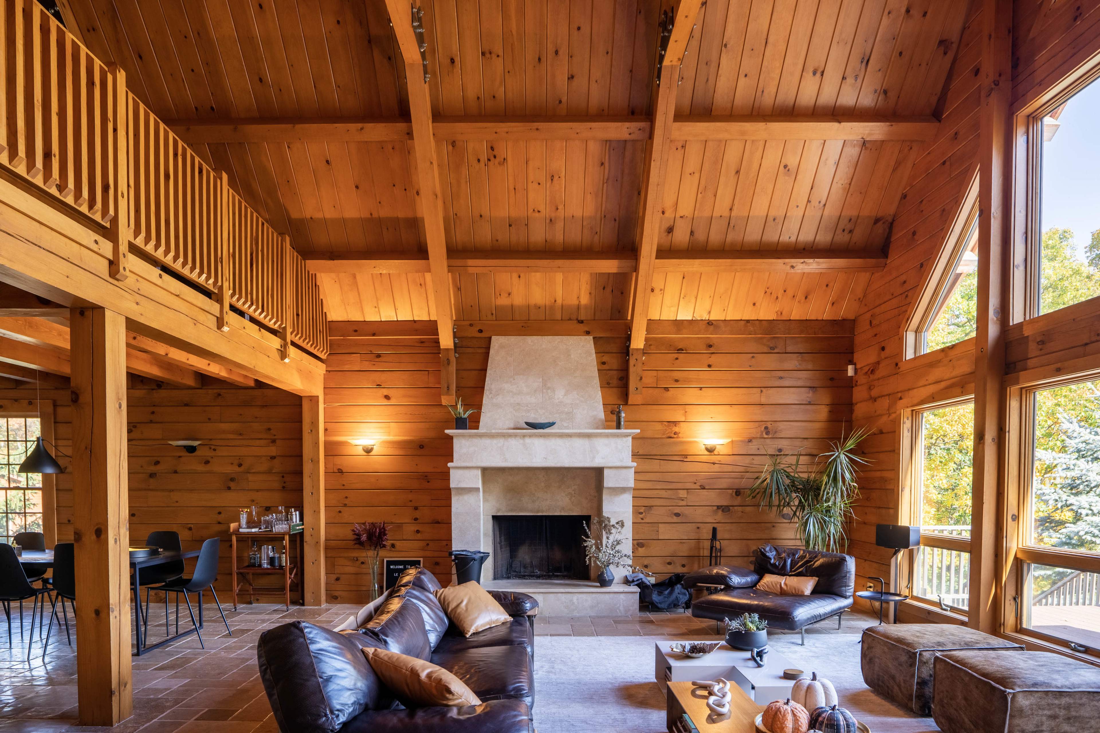 A spacious wooden living room features a high-ceiling design with a prominent stone fireplace and large windows illuminating the space.