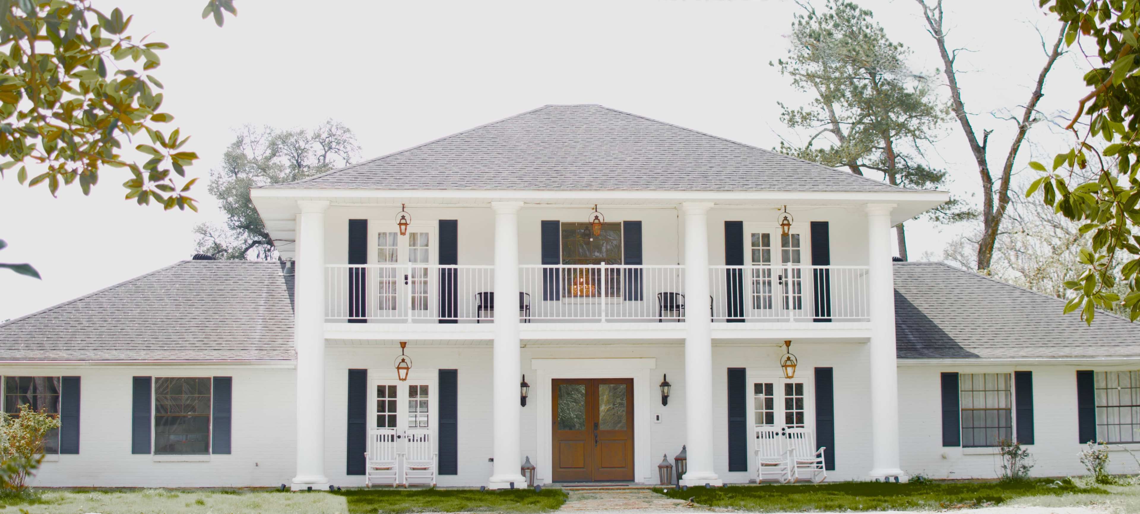 The image shows a large, two-story white house with a central entrance, four columns, and balconies on the upper level.