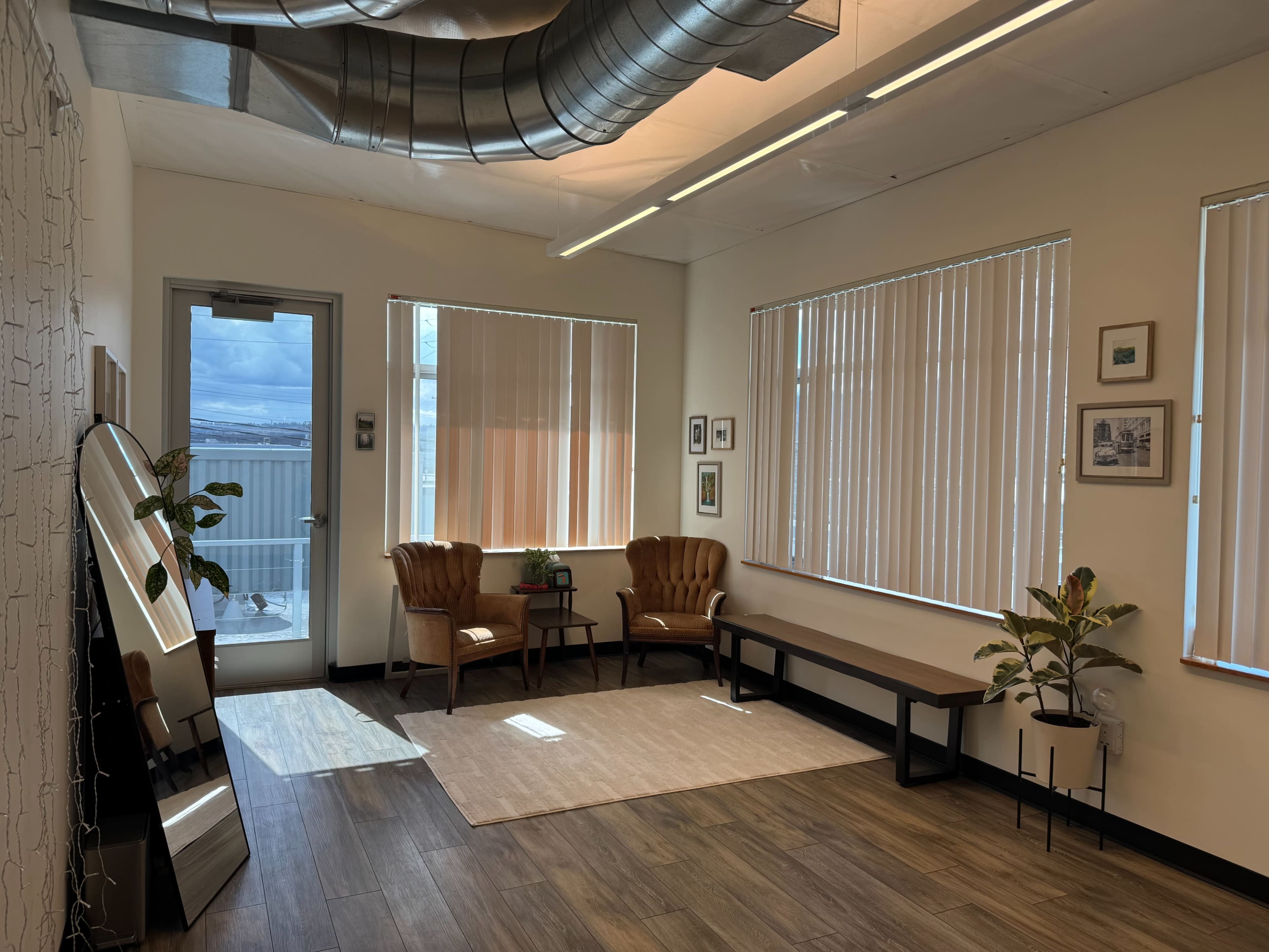 The image shows a bright, sparsely furnished room with two armchairs, a small table, a bench, and large windows with vertical blinds.
