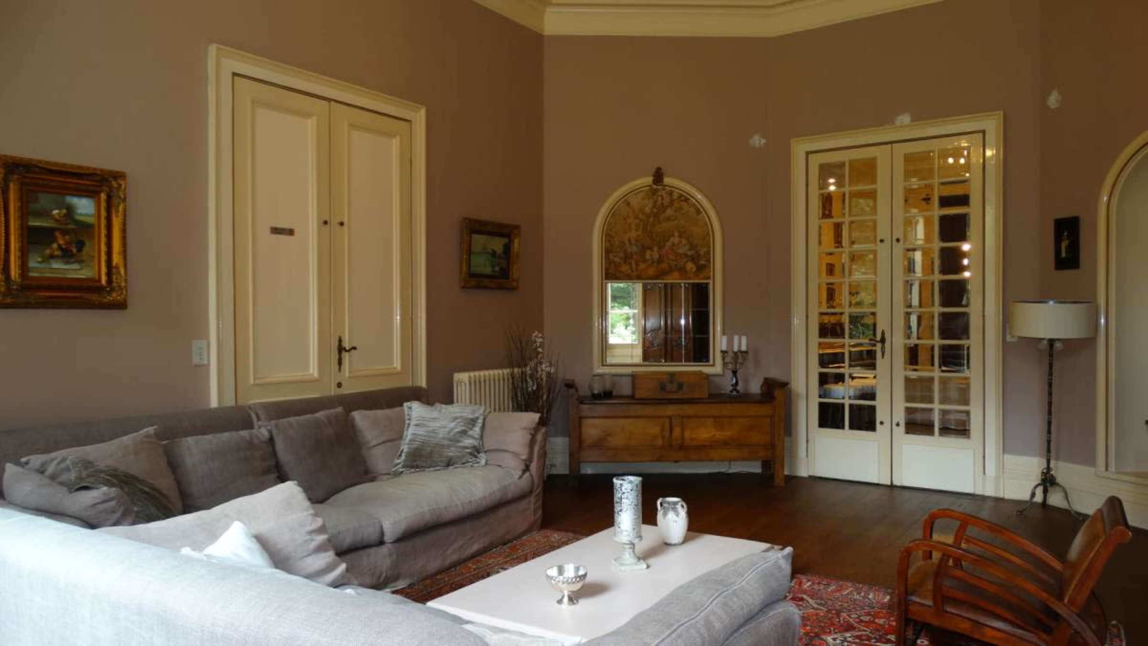 The image depicts a furnished living room with a gray sofa, wooden furniture, and a mirror, set against a pale wall and featuring an arched doorway.