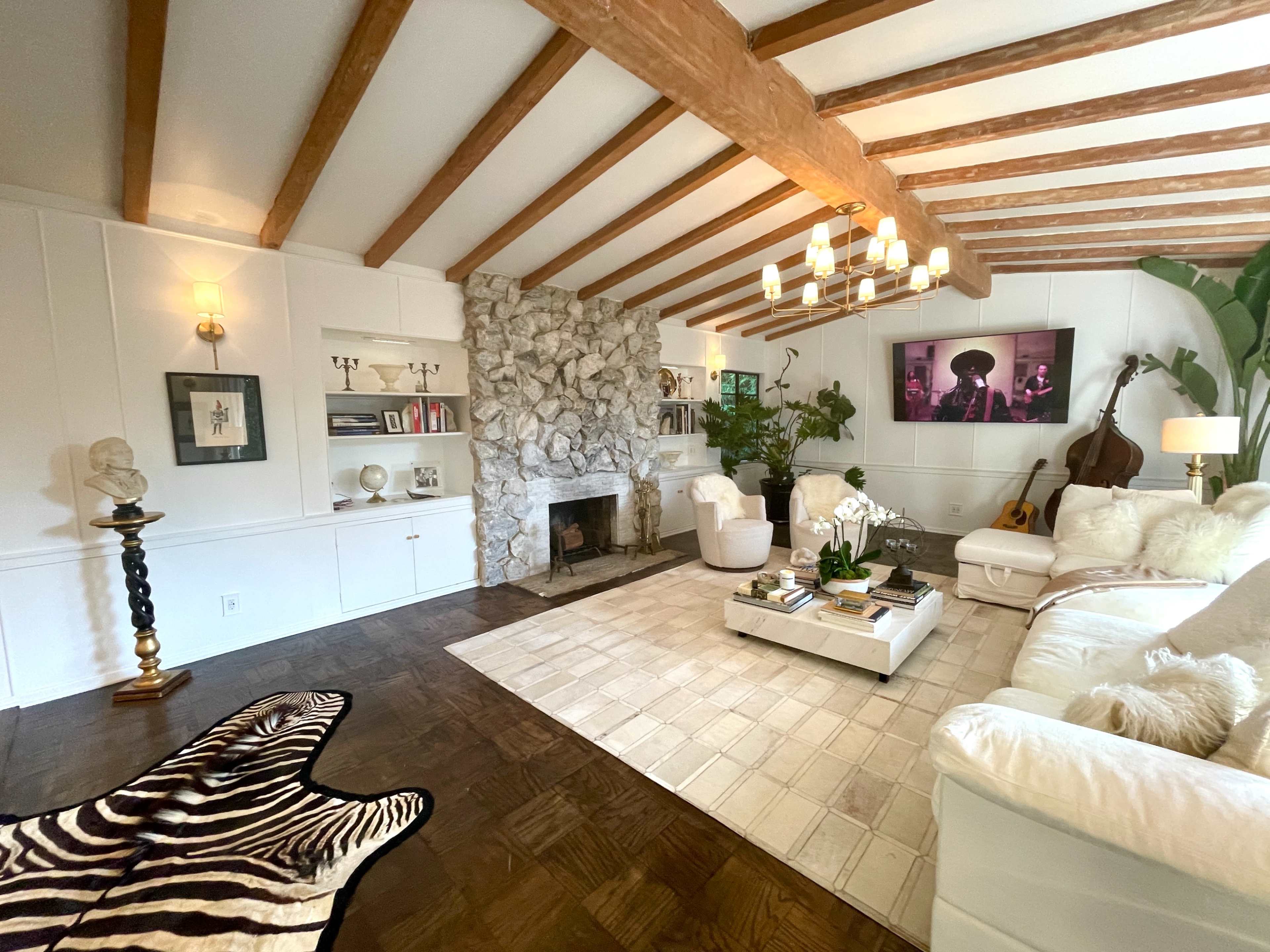 The image depicts a cozy living room with a stone fireplace, wooden beam ceiling, and modern furnishings, including white sofas and a coffee table.