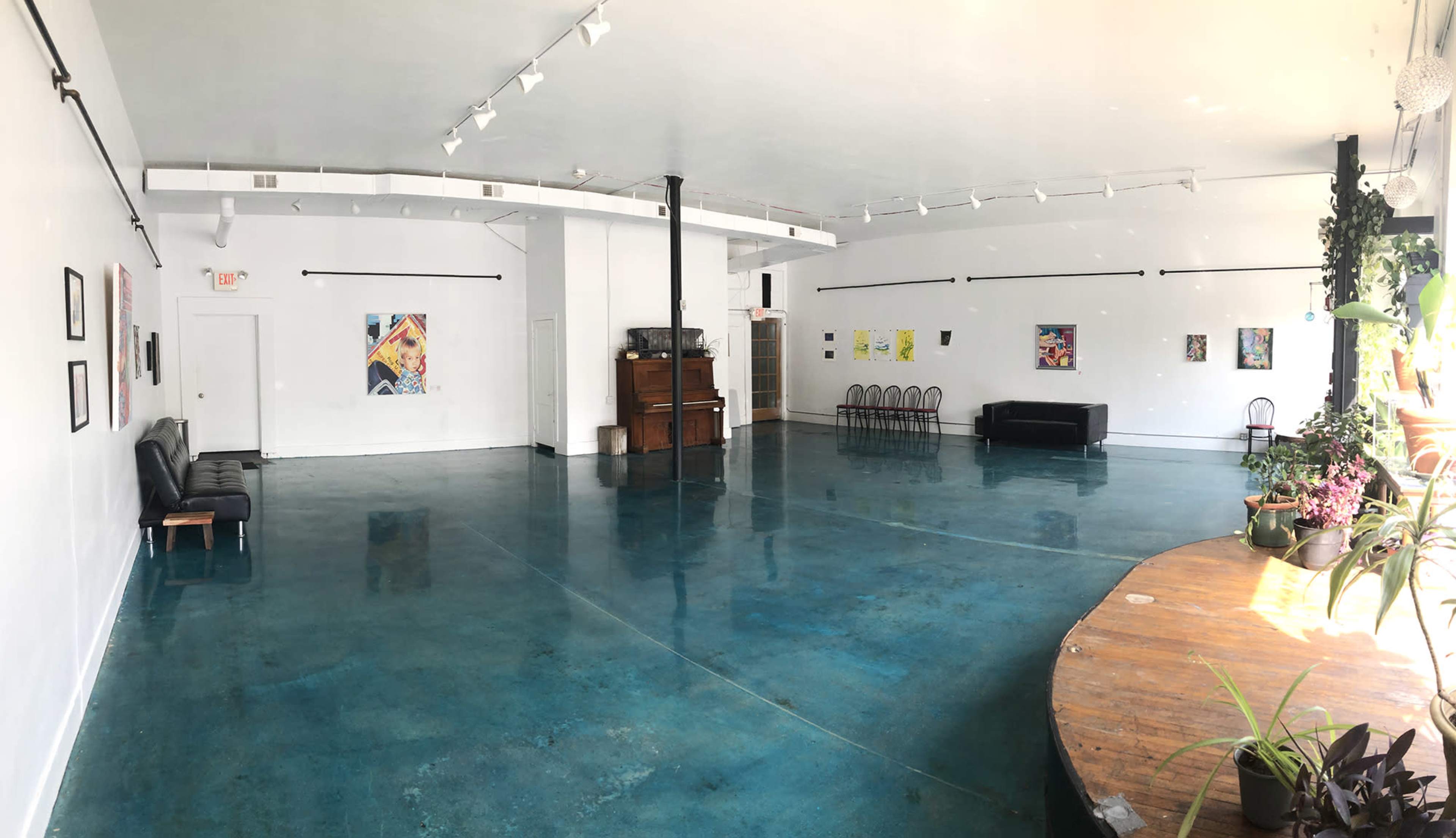 The image shows an empty art gallery with turquoise concrete floors, white walls adorned with colorful artwork, and a circular wooden table surrounded by plants.