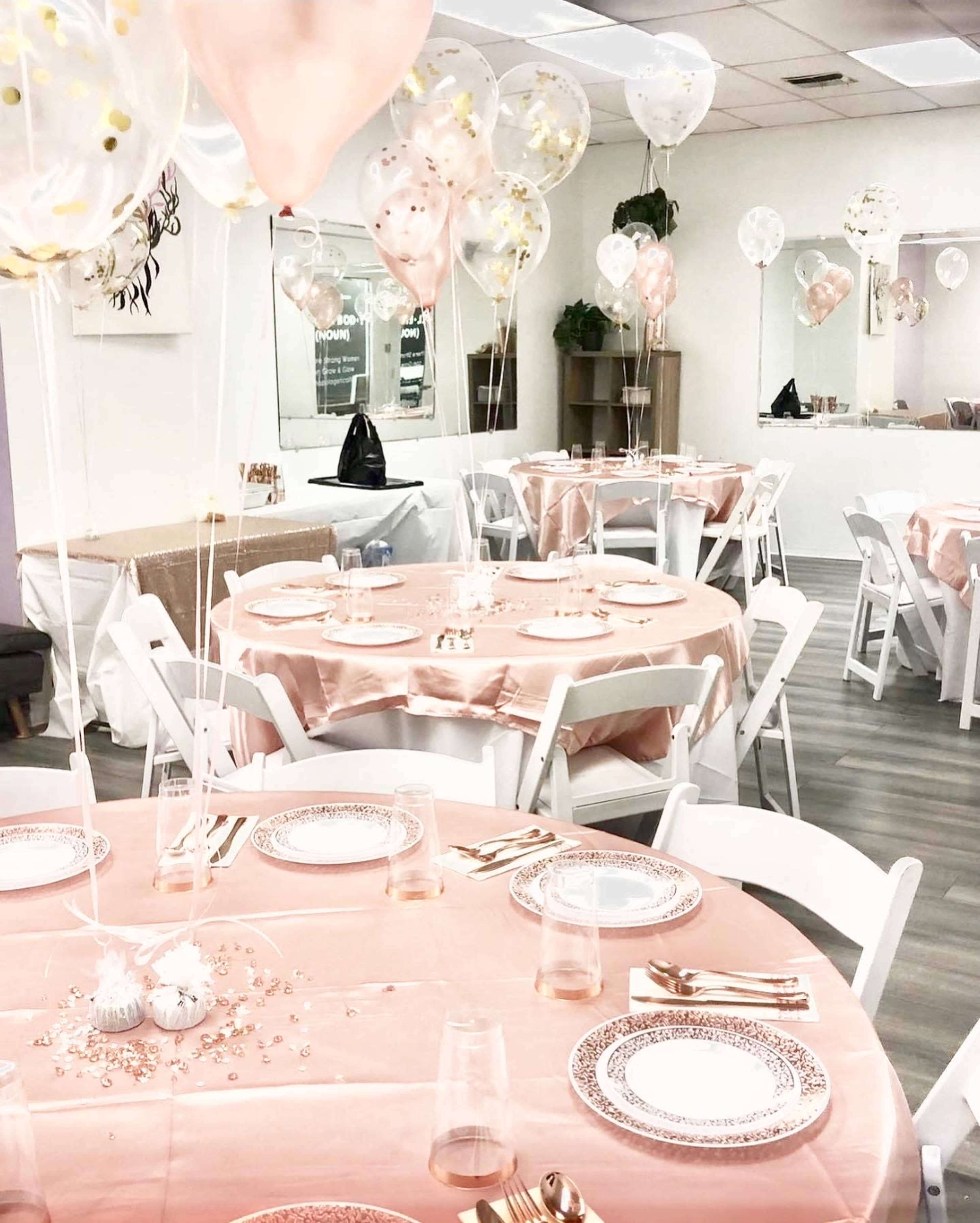 The image shows a decorated event space with round tables draped in pink tablecloths, each set with plates, glasses, and balloons.