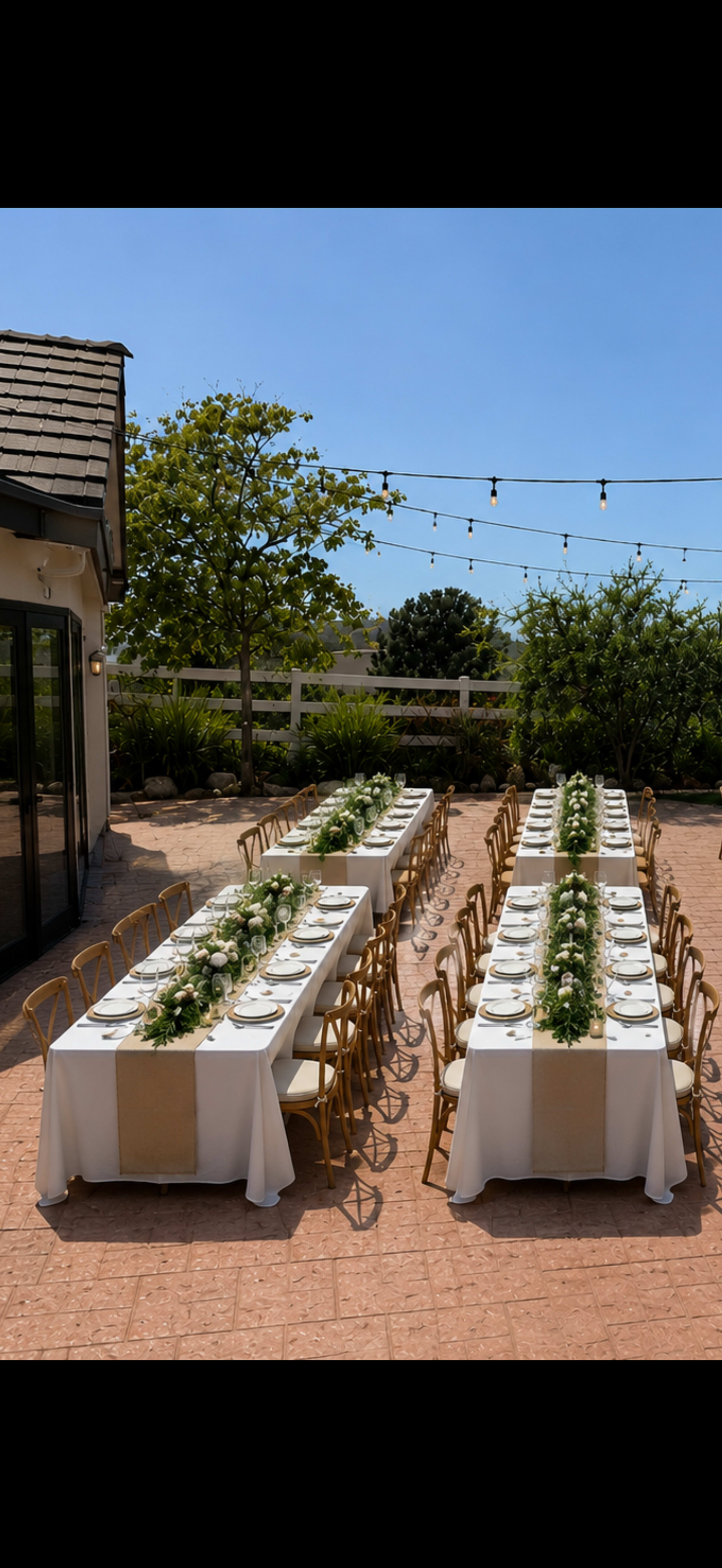 Two long tables are set for a meal, adorned with white tablecloths and centerpieces, arranged outdoors under clear blue skies.