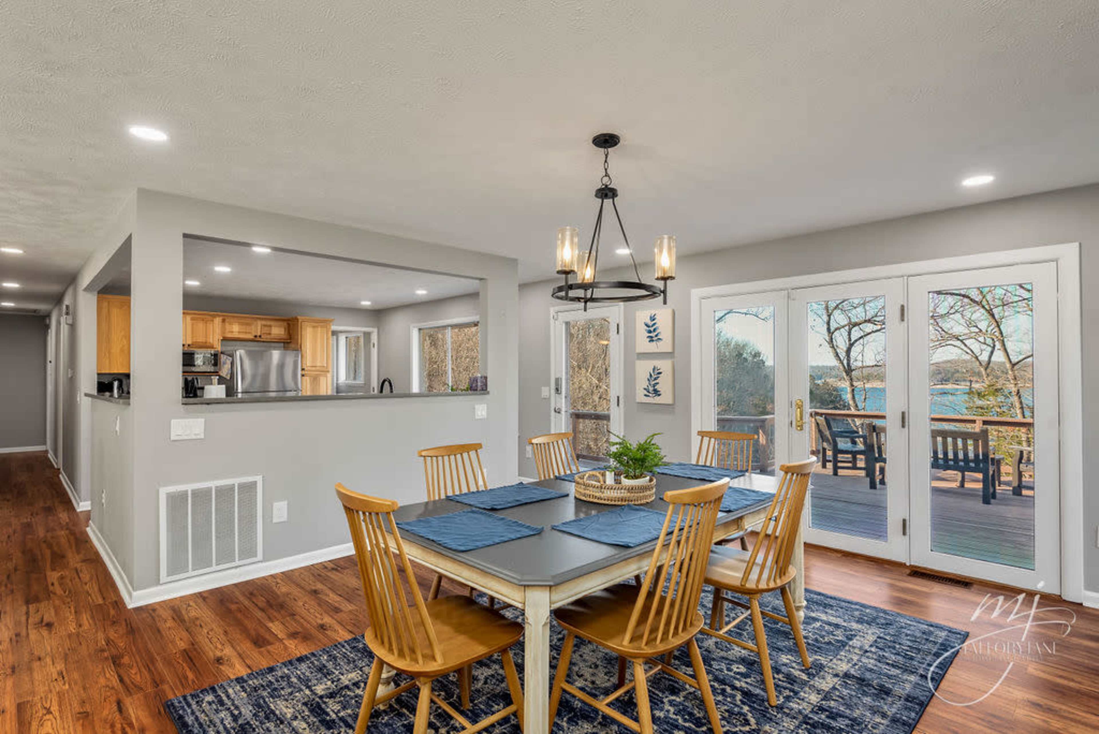 A dining area features a light-colored table with wooden chairs, set against a backdrop of large windows and a view of the outdoors.