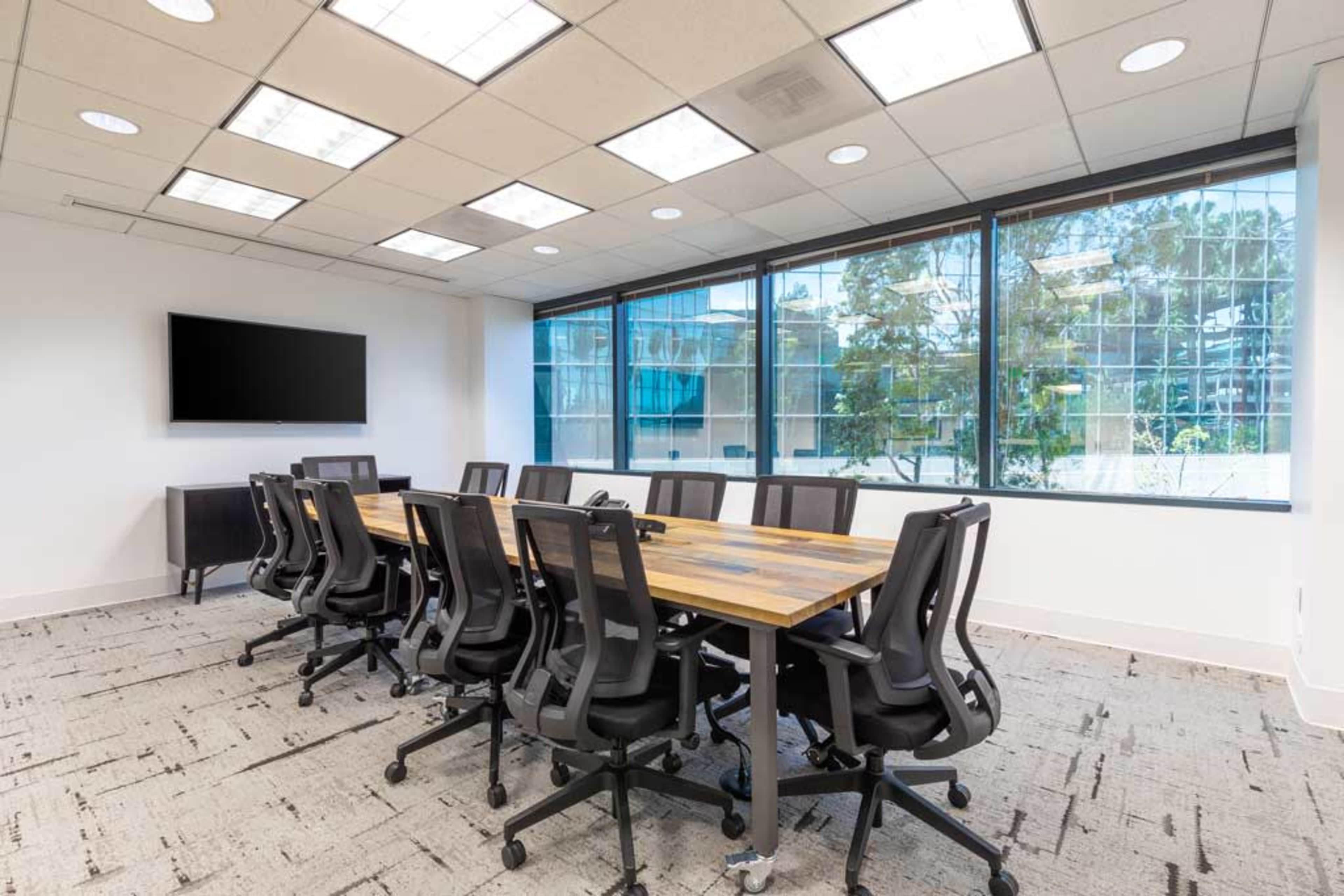 A modern meeting room features a large wooden table surrounded by ergonomic chairs, with large windows providing an outdoor view and a wall-mounted screen.