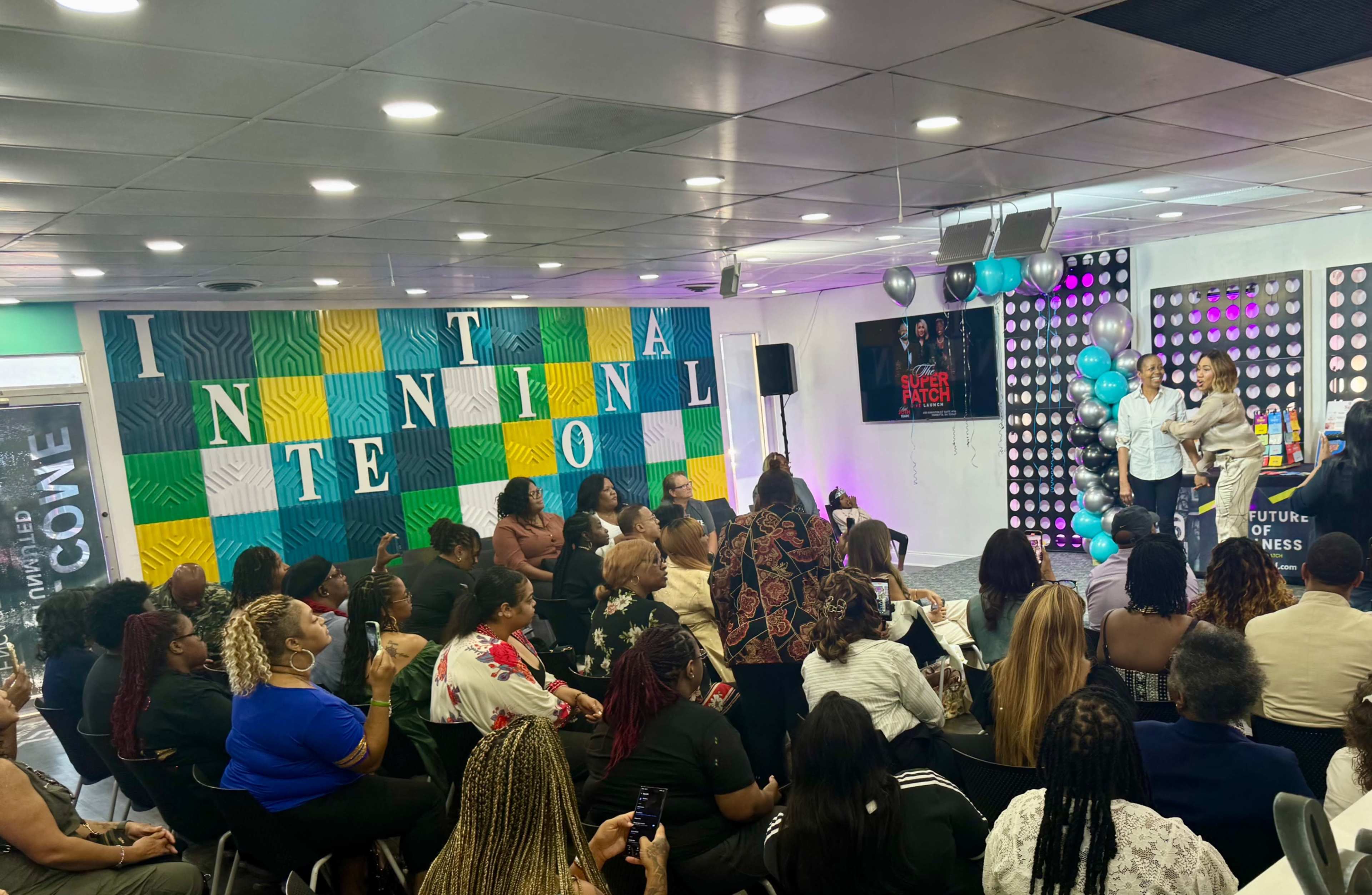 The image shows a large group of people seated in an indoor event space, with colorful wall decorations and a stage where two women are speaking to the audience.
