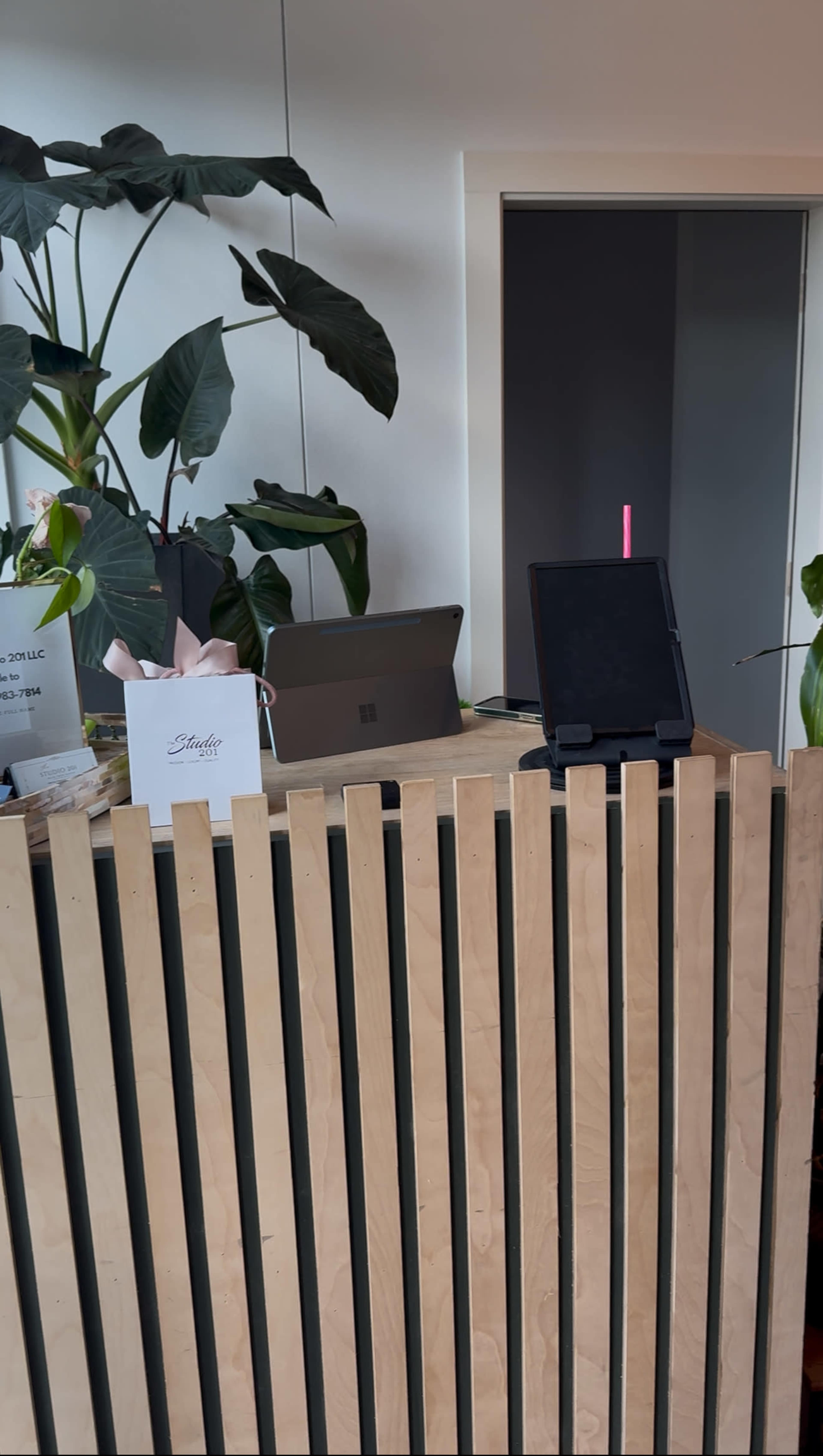 The image shows a reception area with a wooden slatted counter, a tablet on a stand, a laptop, and several green plants.