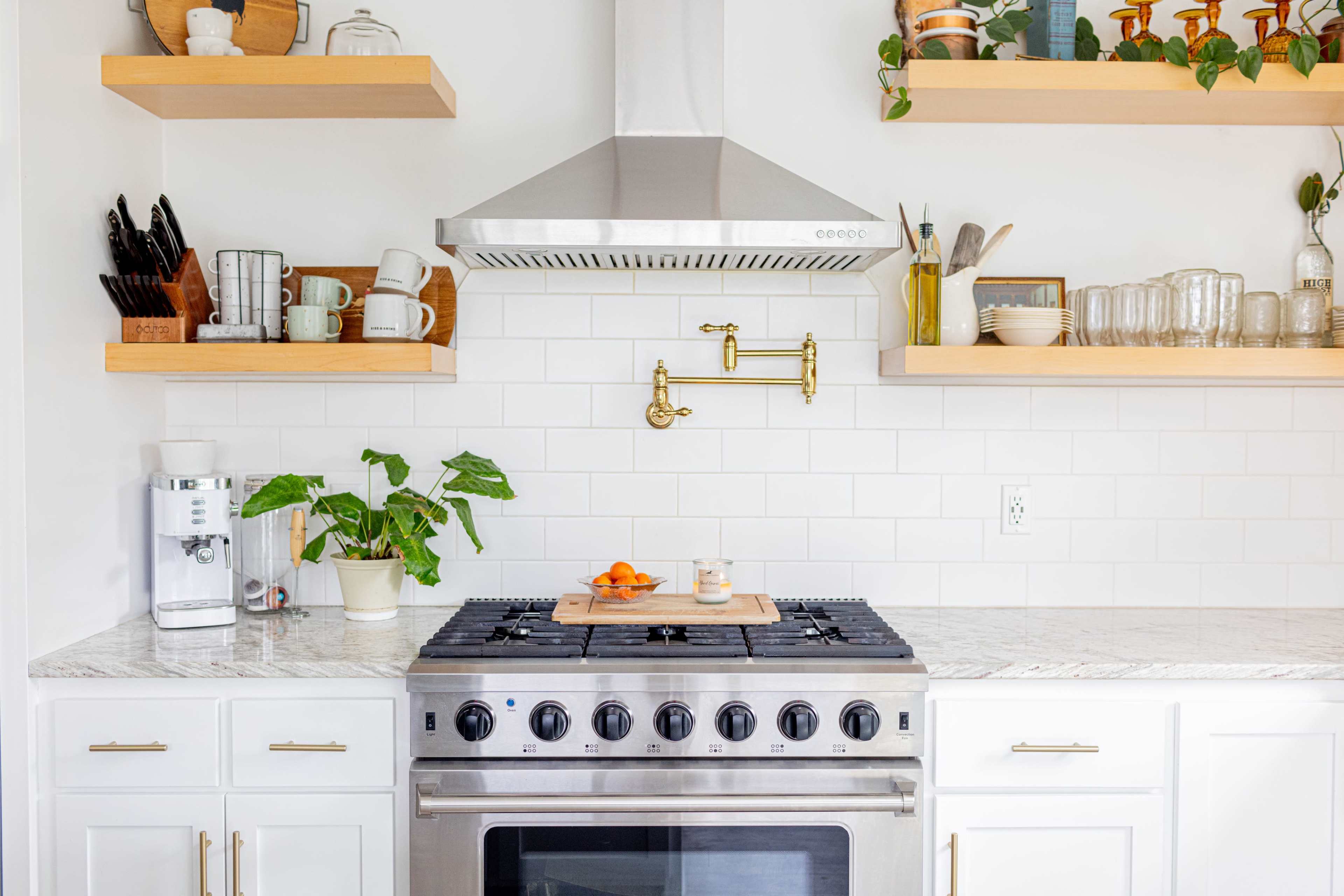 The image features a modern kitchen with a stainless steel gas stove, a gold wall-mounted faucet, open shelving displaying dishes and plants, and a marble countertop.