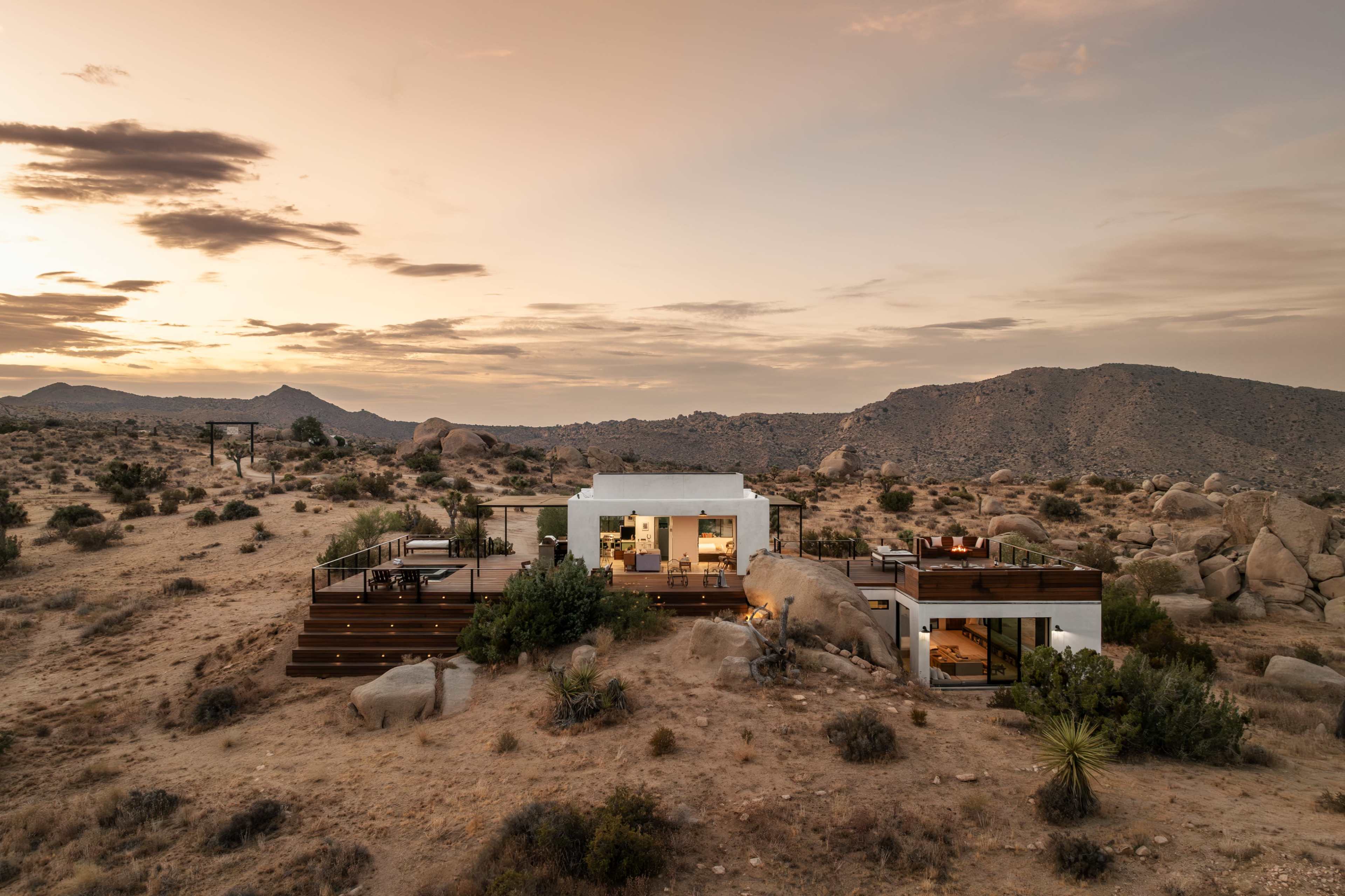 A modern house is surrounded by a desert landscape, featuring rocky terrain and distant mountains under a sunset sky.
