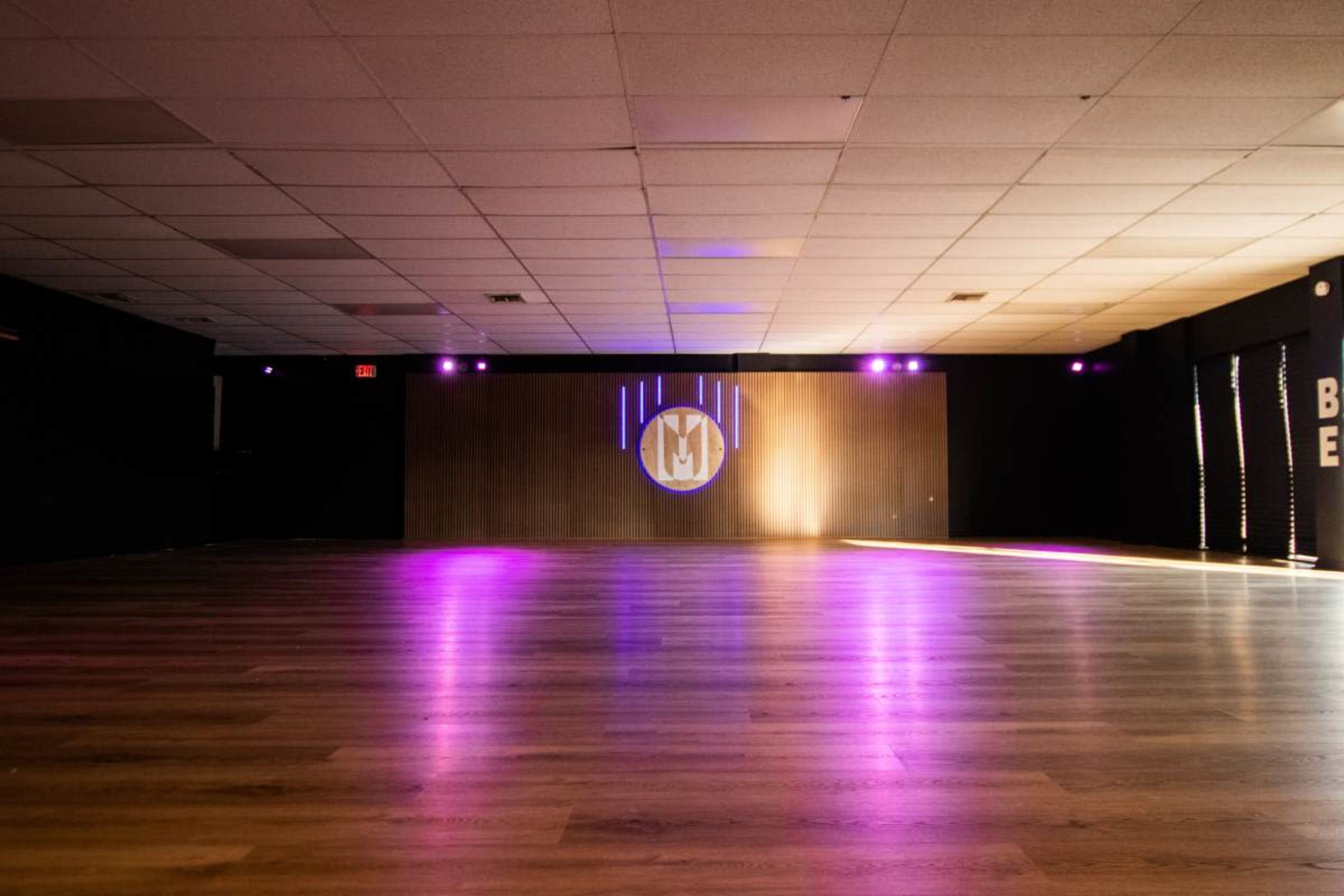 An empty indoor space with polished wooden floors features a projection of a logo on the wall, illuminated by purple lights.