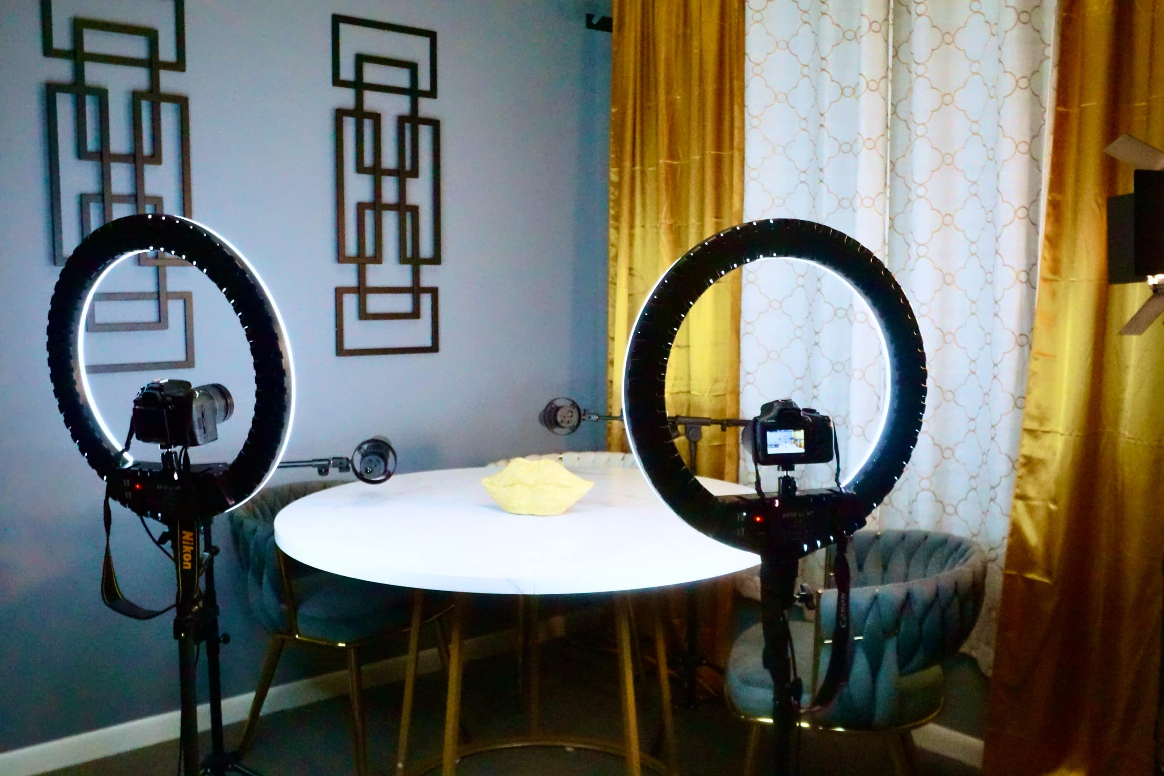 The image shows a well-lit setup featuring two ring lights positioned on either side of a round white table with a decorative bowl in the center, set against a backdrop of patterned curtains and geometric wall art.