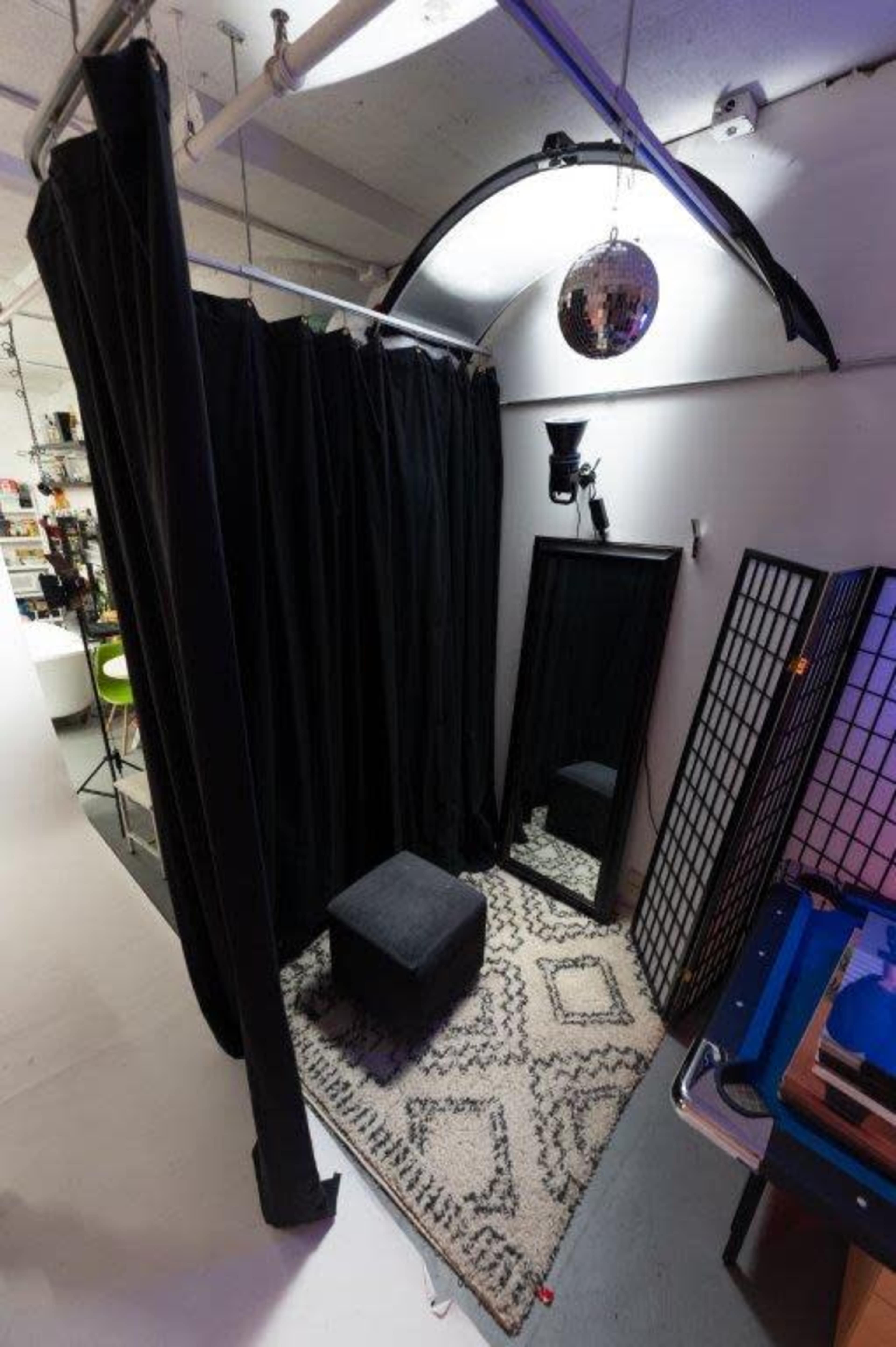 The image shows a small room with black curtains, a mirror, a black stool, a patterned rug, and a disco ball hanging from the ceiling.