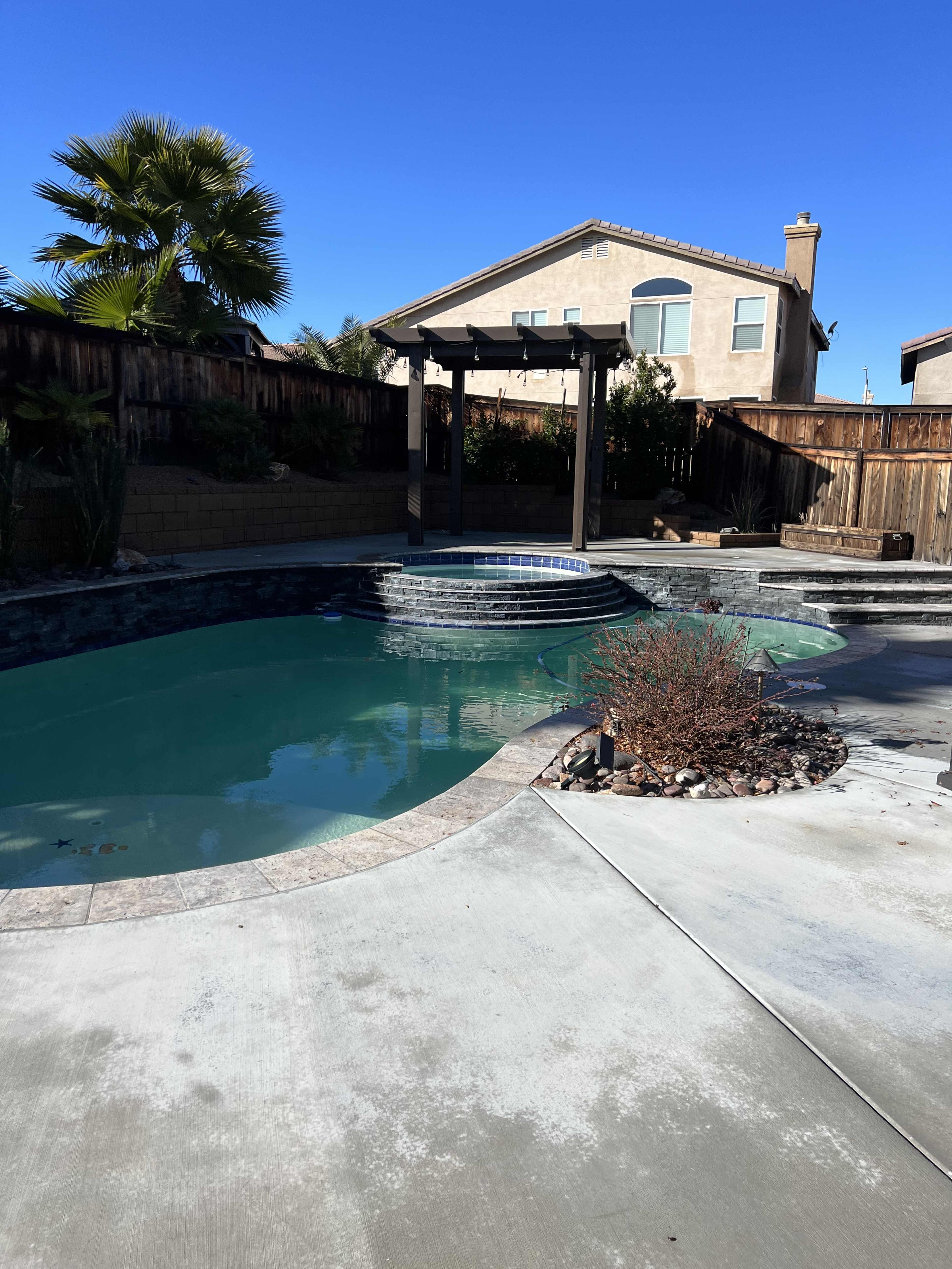 A landscaped backyard with a swimming pool, a stone waterfall feature, and a pergola, adjacent to a two-story house.