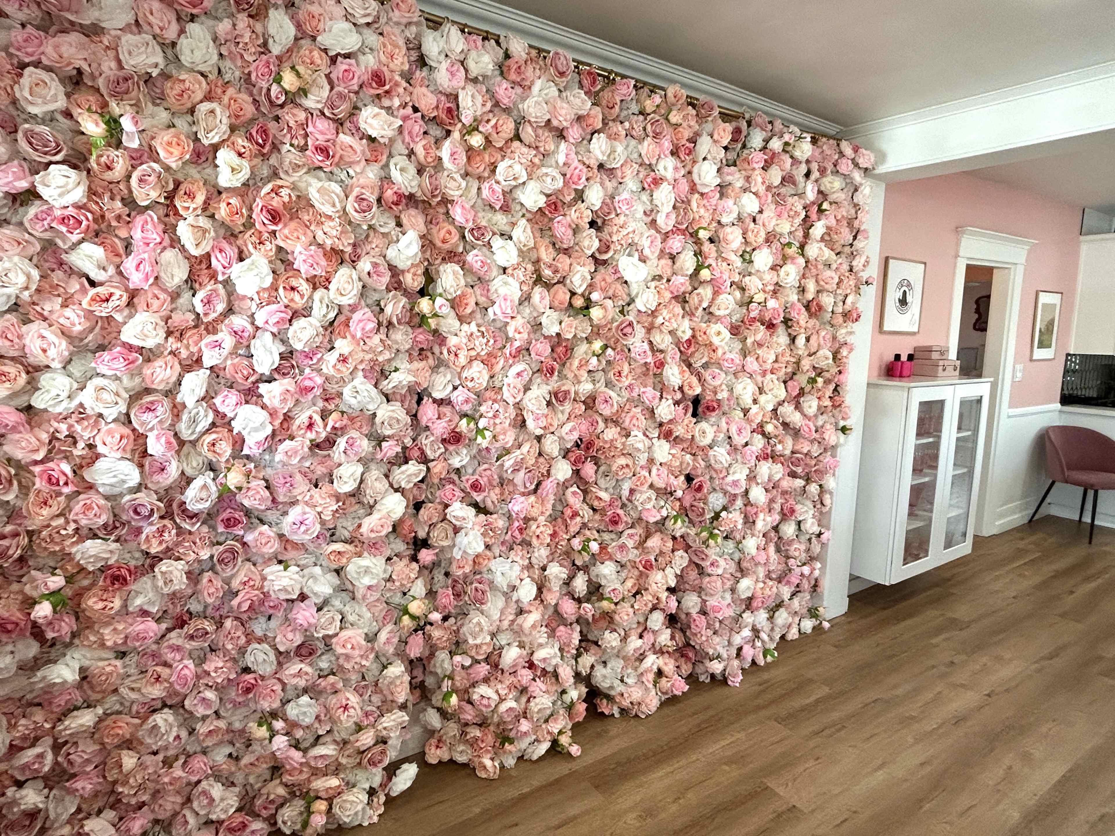 The image shows a wall covered with a dense arrangement of pink and white artificial roses, creating a floral backdrop in a room with wooden flooring and minimal furnishings.