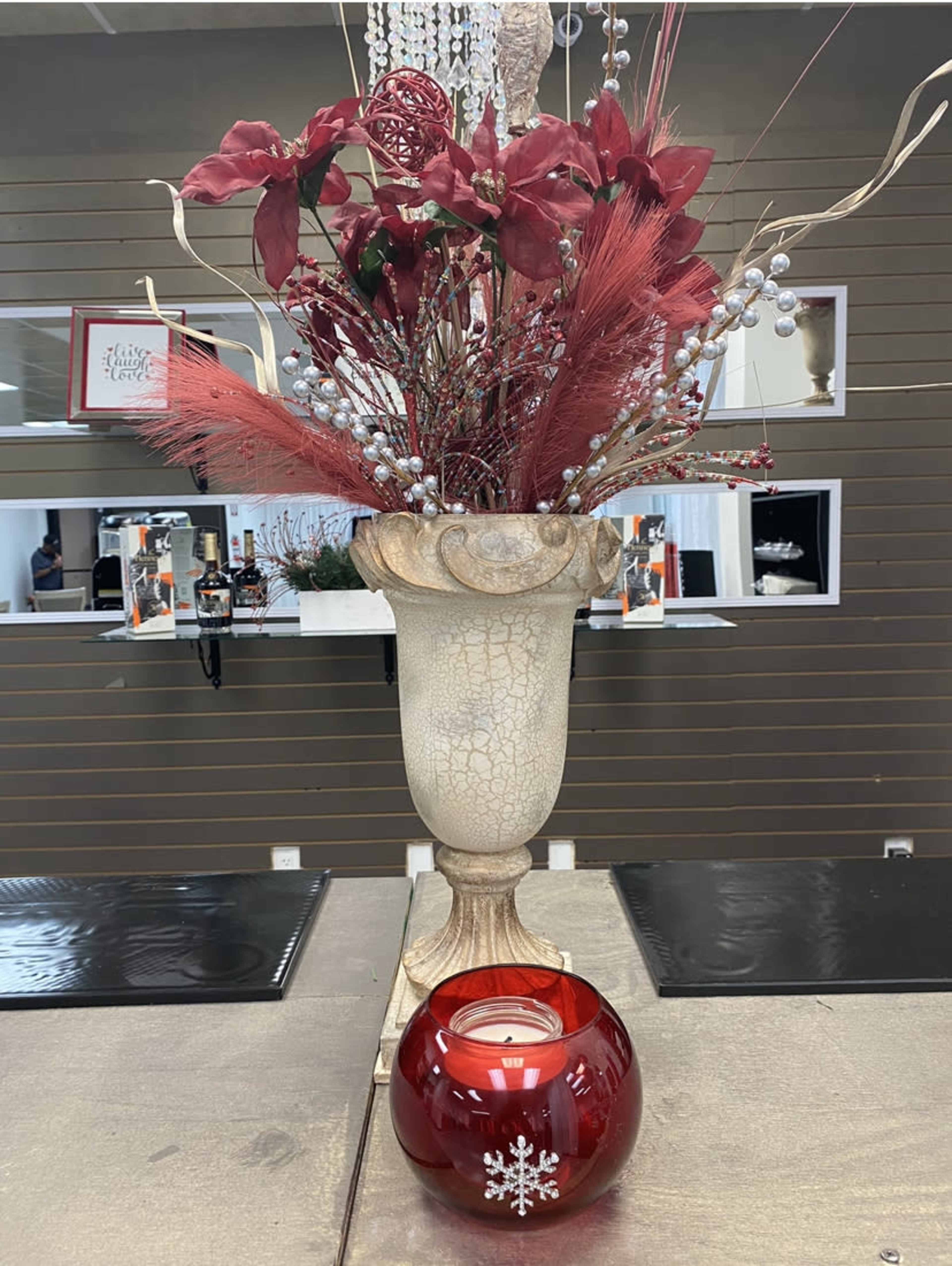 A decorative vase filled with red flowers and foliage stands next to a red candle holder with a snowflake design on a table.