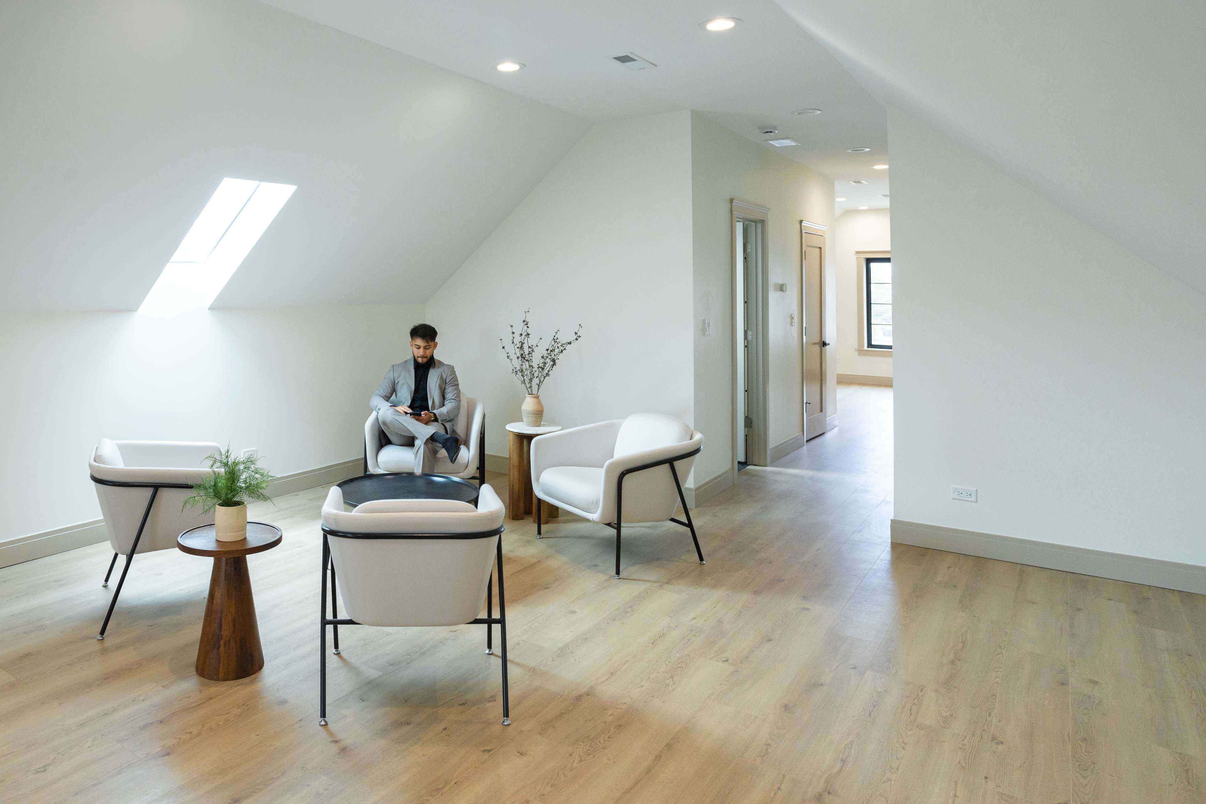 A man sits in a modern, light-filled room furnished with two chairs and a small table, with an open hallway visible in the background.