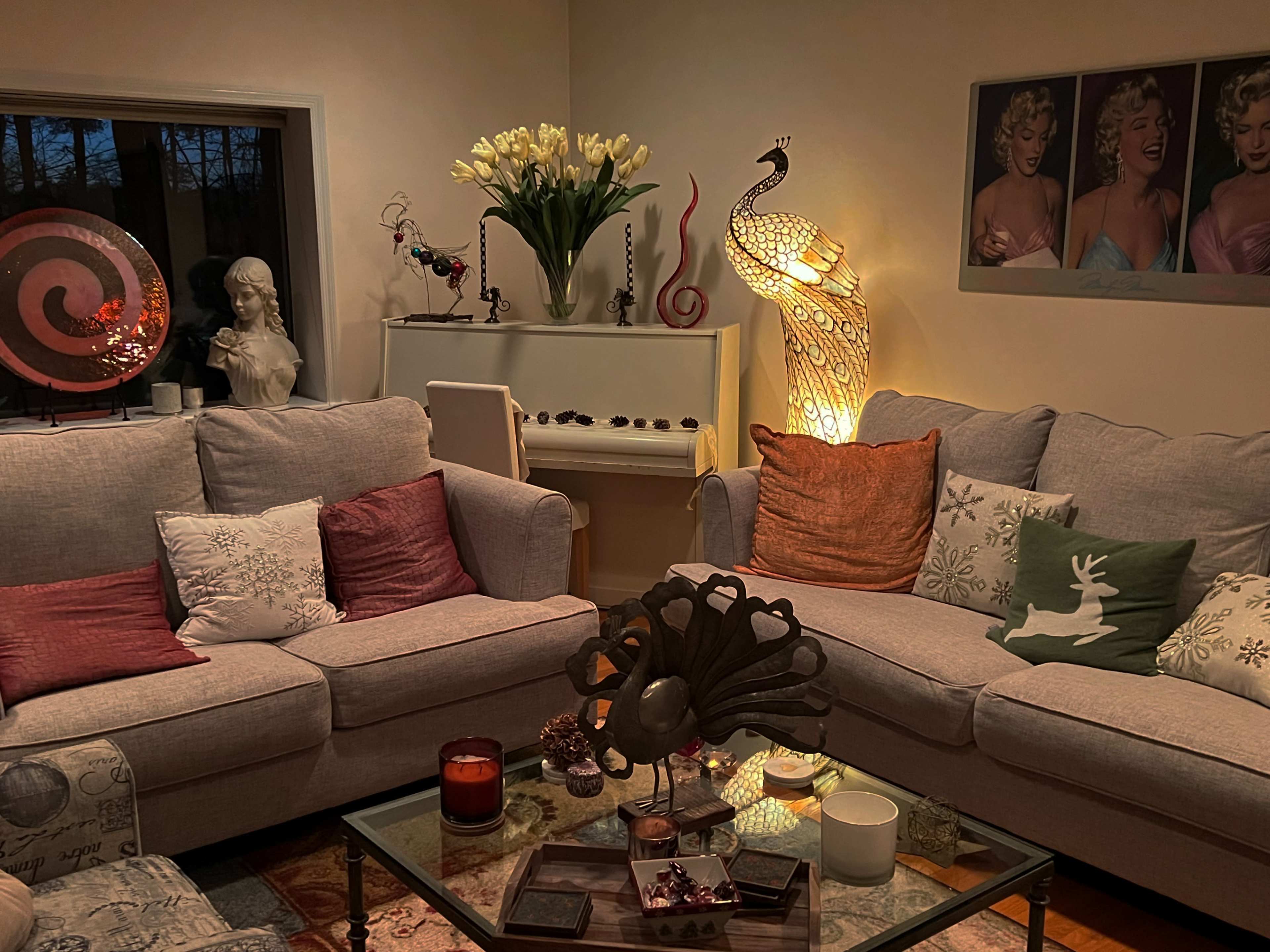 The scene features a cozy living room with a light-colored sectional sofa adorned with decorative pillows, a peacock lamp, and a white piano in the corner, surrounded by various decorative items and artwork.