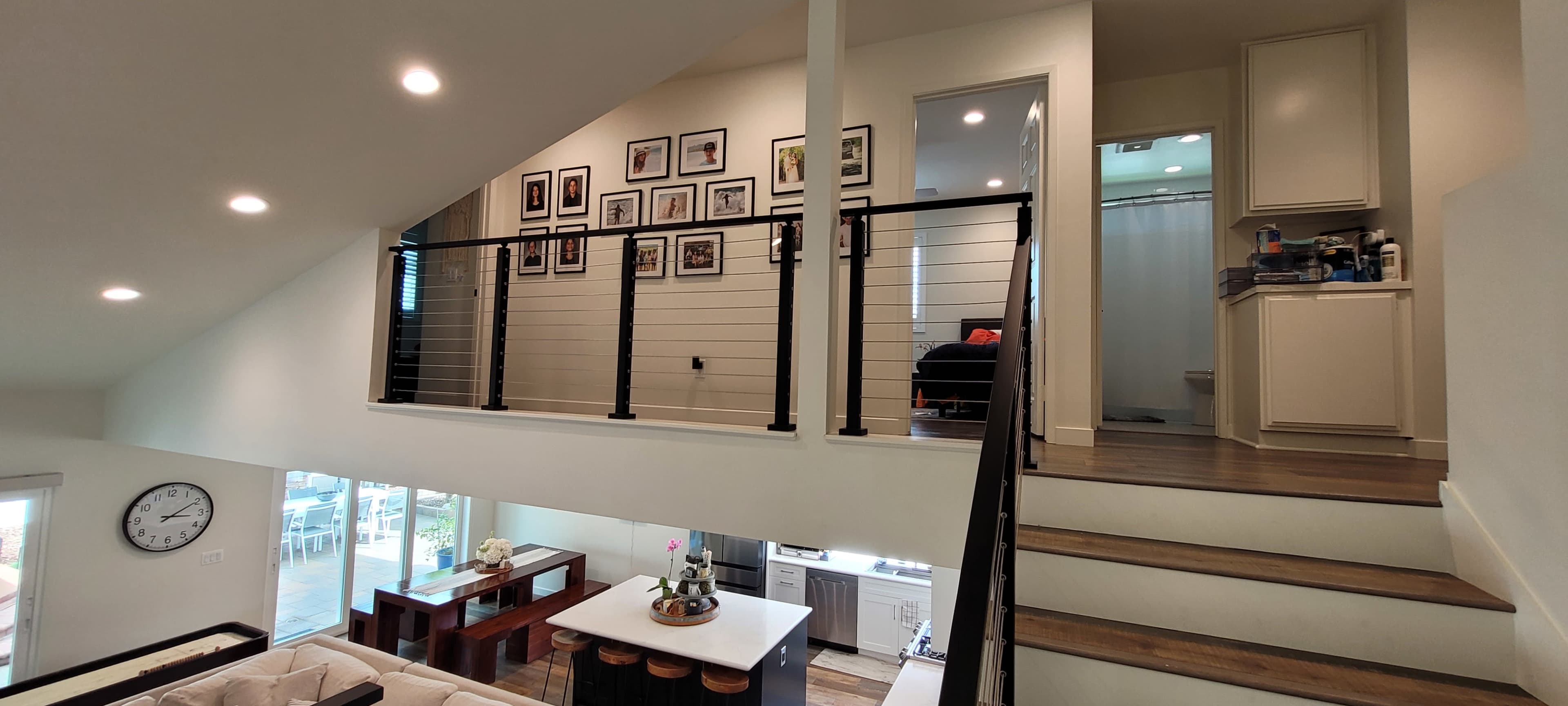 The image shows a modern interior of a two-story home, featuring an open stairway, a gallery wall with framed photos, and a view of a bathroom and bedroom at the top of the stairs.