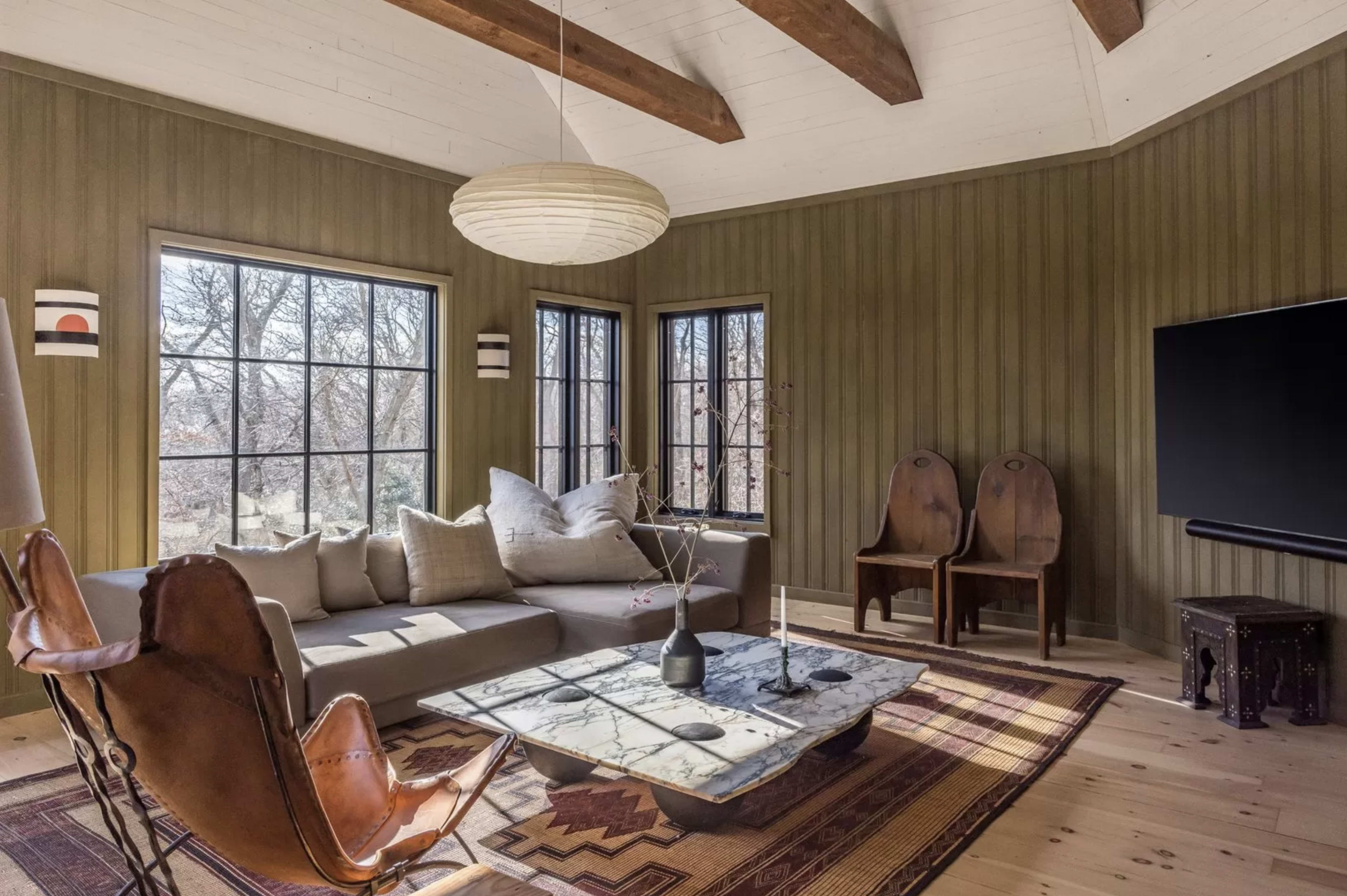 A cozy living room with a sectional sofa, a coffee table, a patterned rug, and two wooden chairs against a wall of windows.