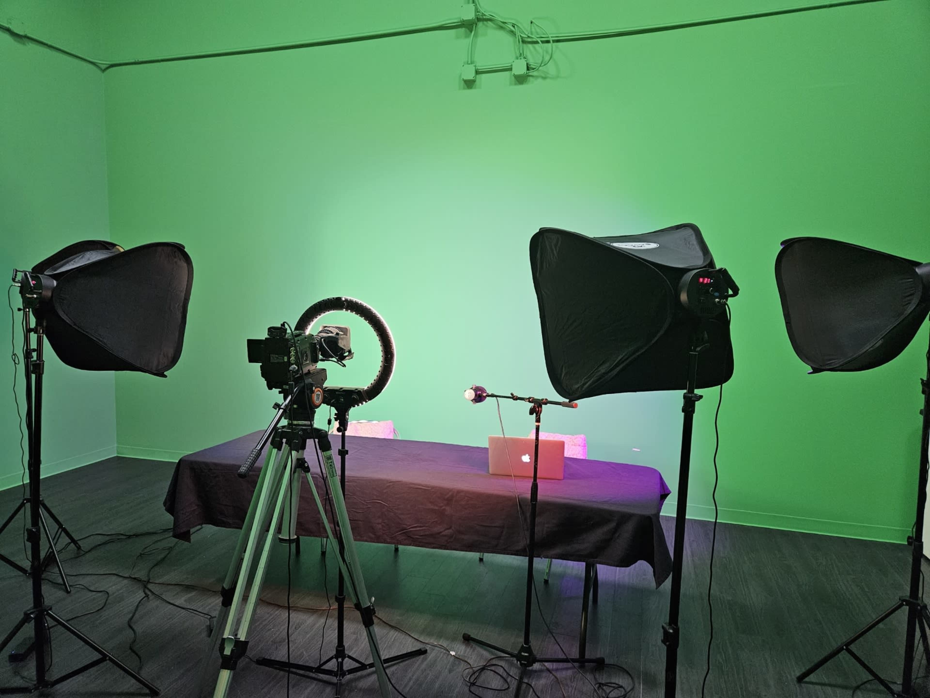 A video recording setup featuring a table with a laptop, a camera on a tripod, softbox lights, and a ring light in front of a green background.