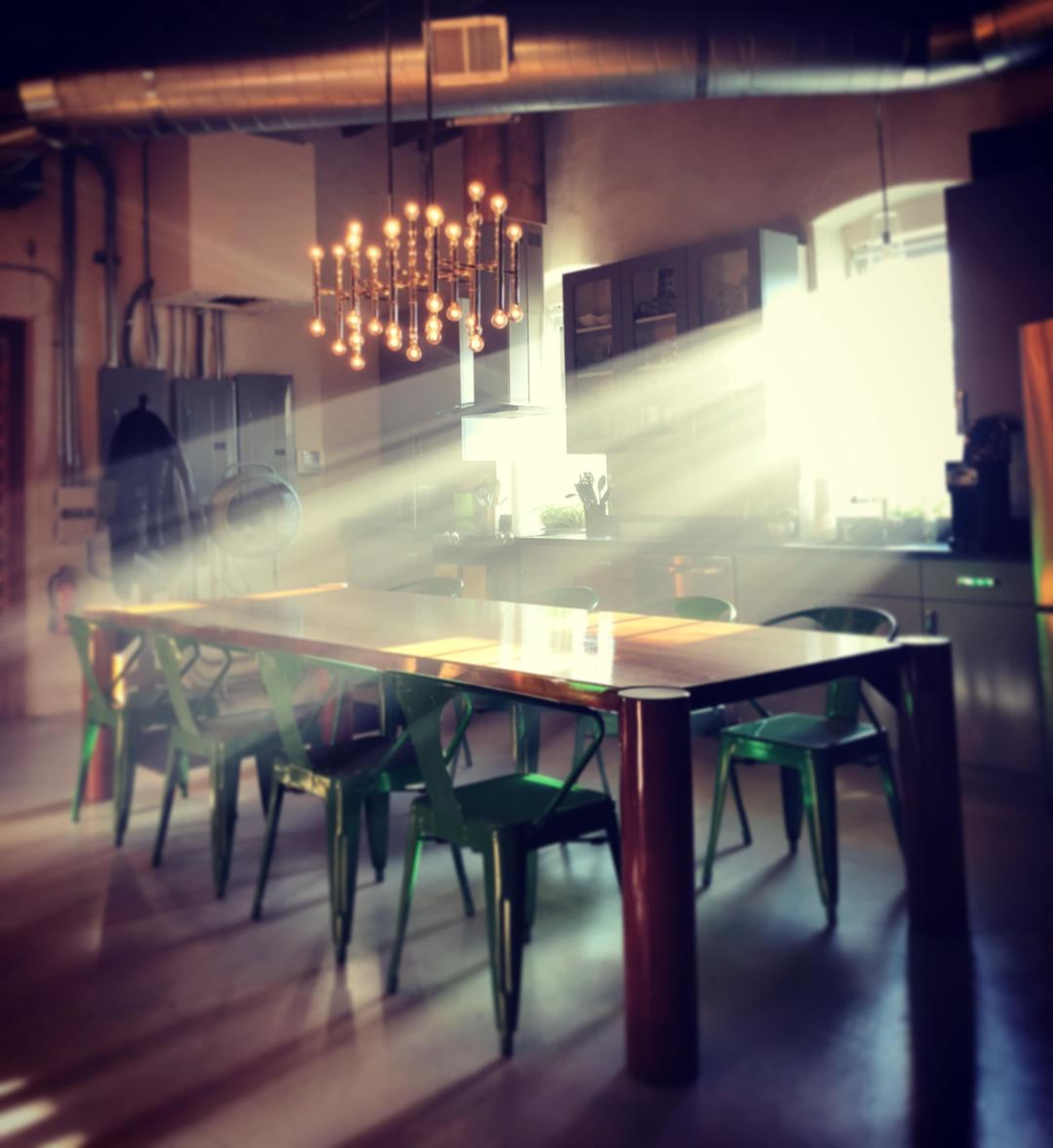 A large wooden dining table surrounded by green chairs is illuminated by sunlight streaming through a window in a modern kitchen.