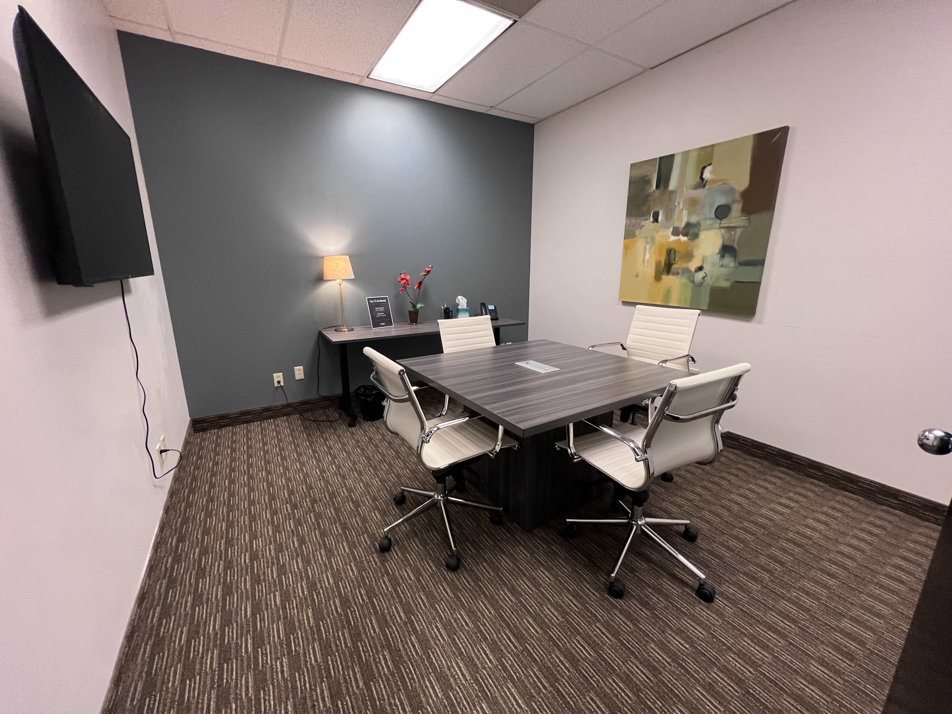 A small conference room with a rectangular table, two white chairs, a desk with a lamp, and an abstract painting on the wall.