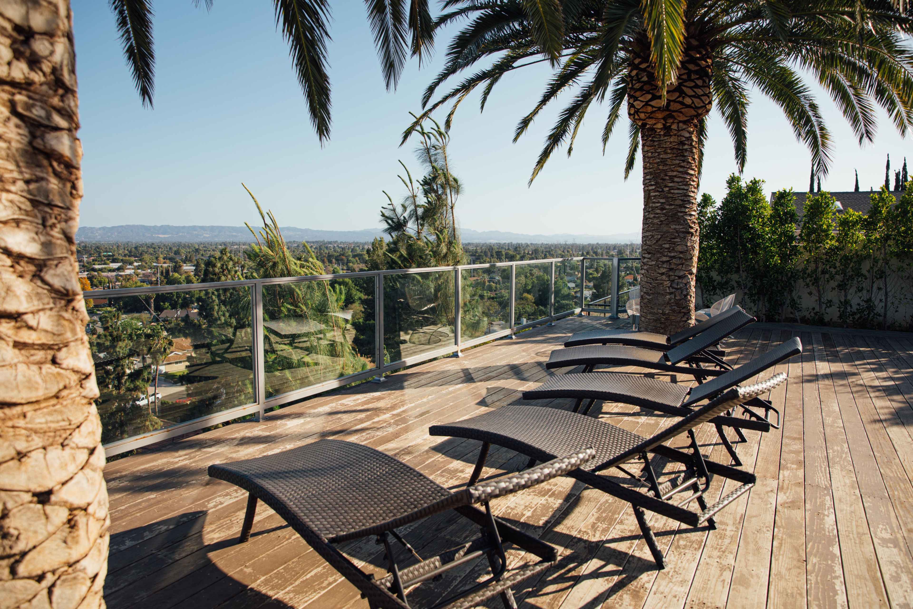 A wooden deck features several black lounge chairs under palm trees with a view of a distant landscape.