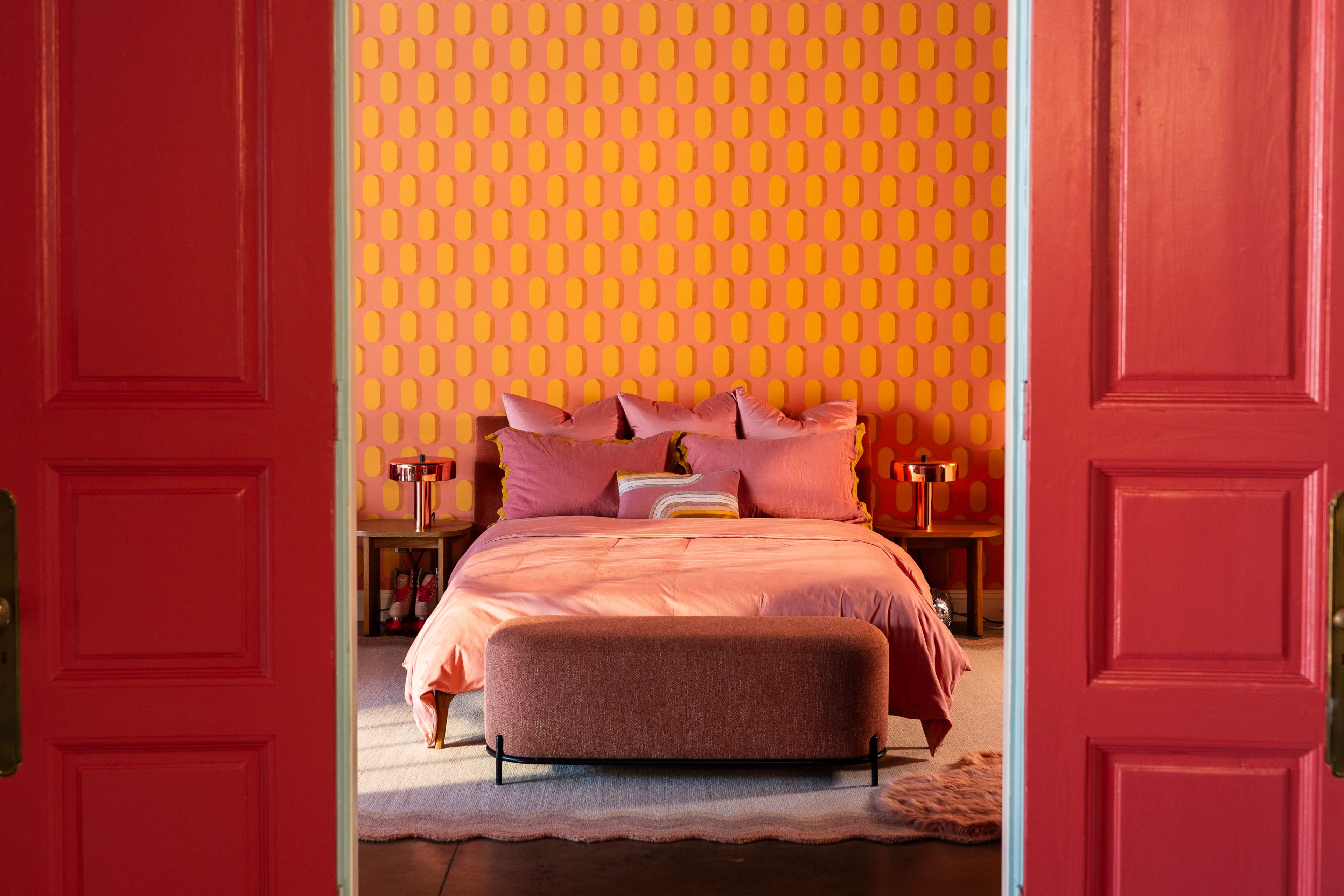 A well-decorated bedroom features a pink bedspread, matching pillows, and a decorative wall with an orange and yellow pattern, framed by red double doors.