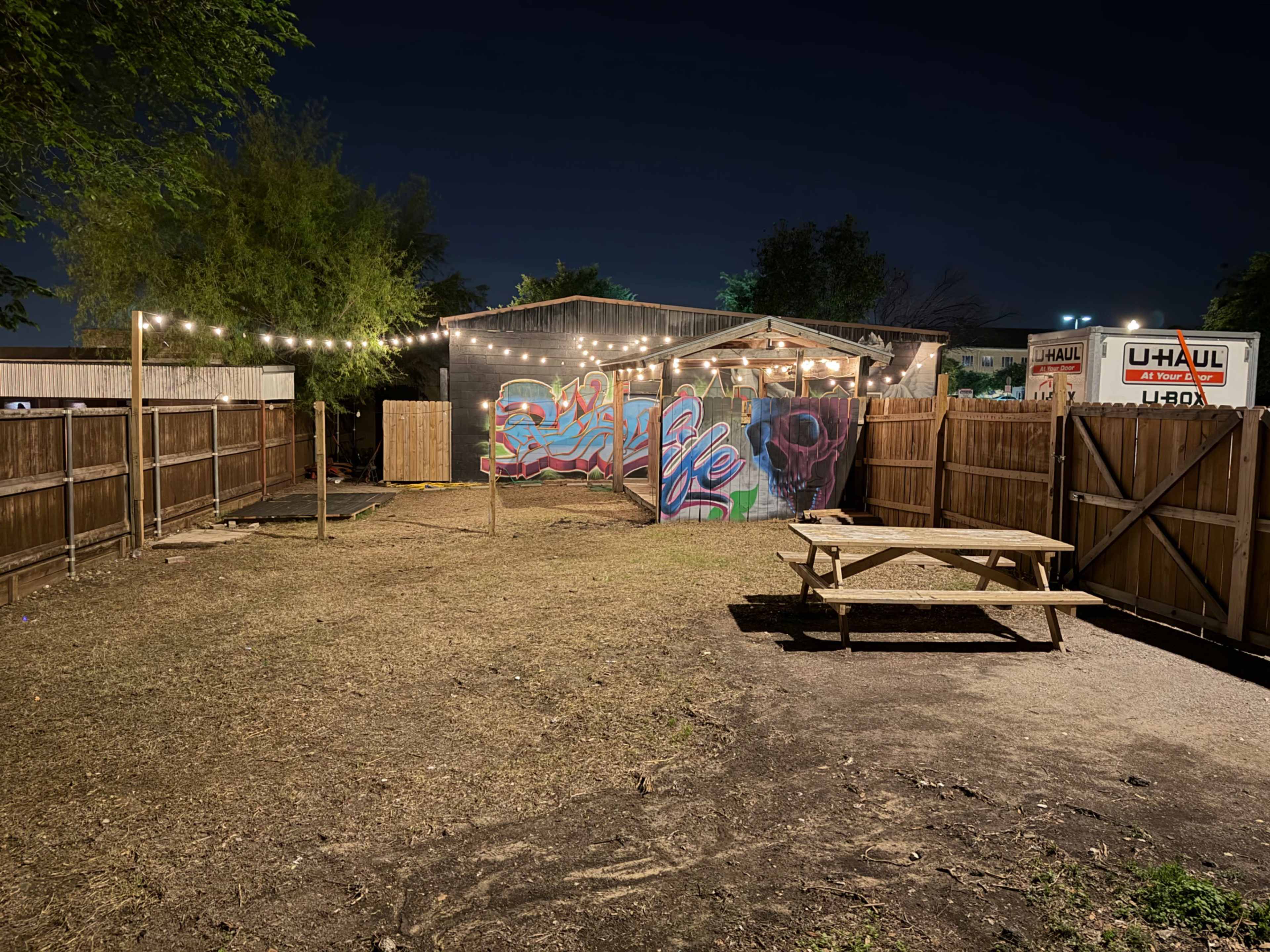 A fenced outdoor space at night features a picnic table, string lights overhead, and a colorful mural on the back wall of a building.