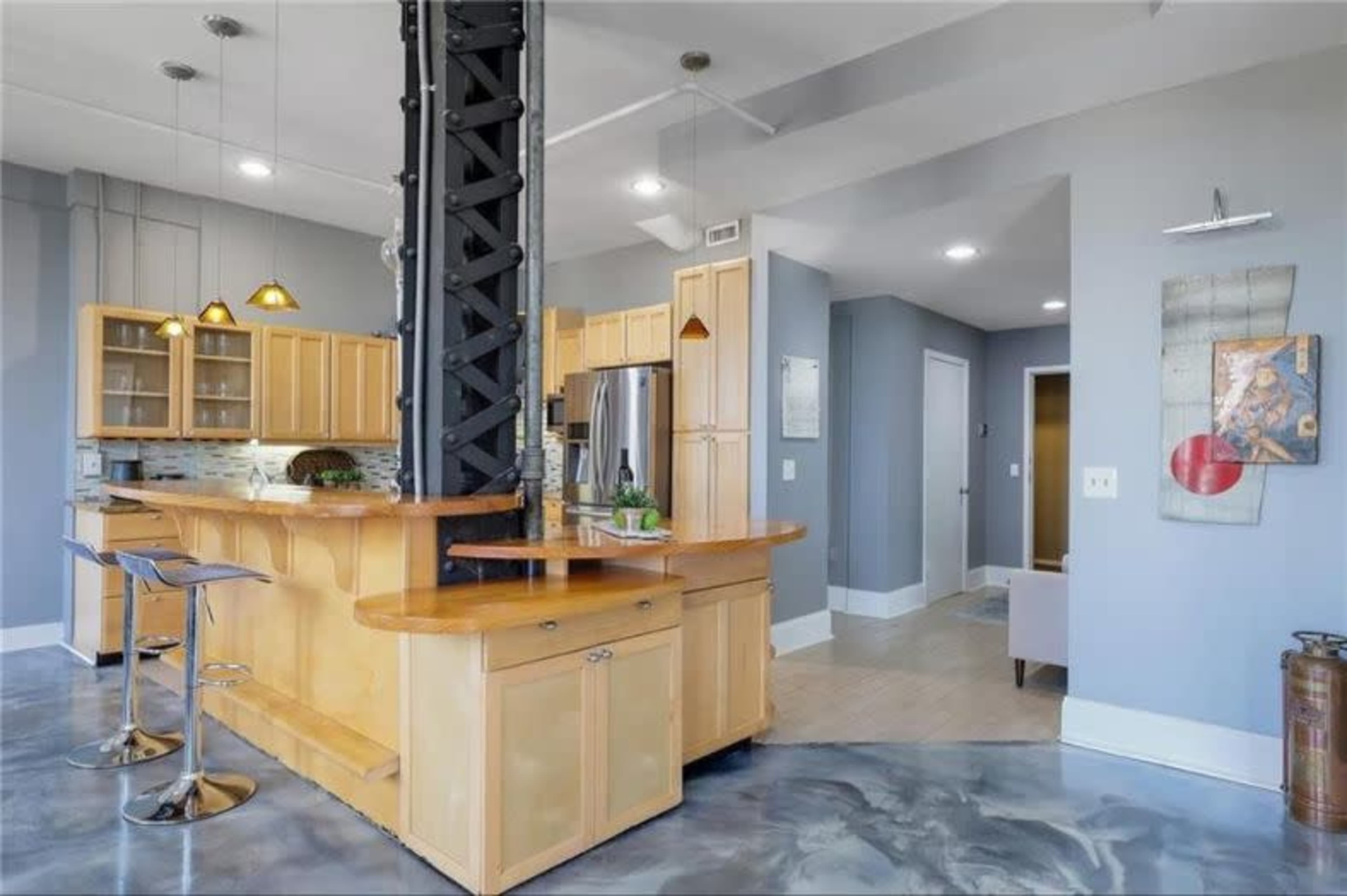 The image shows a modern kitchen with a wooden island and metal support column, featuring bar stools and stainless steel appliances.