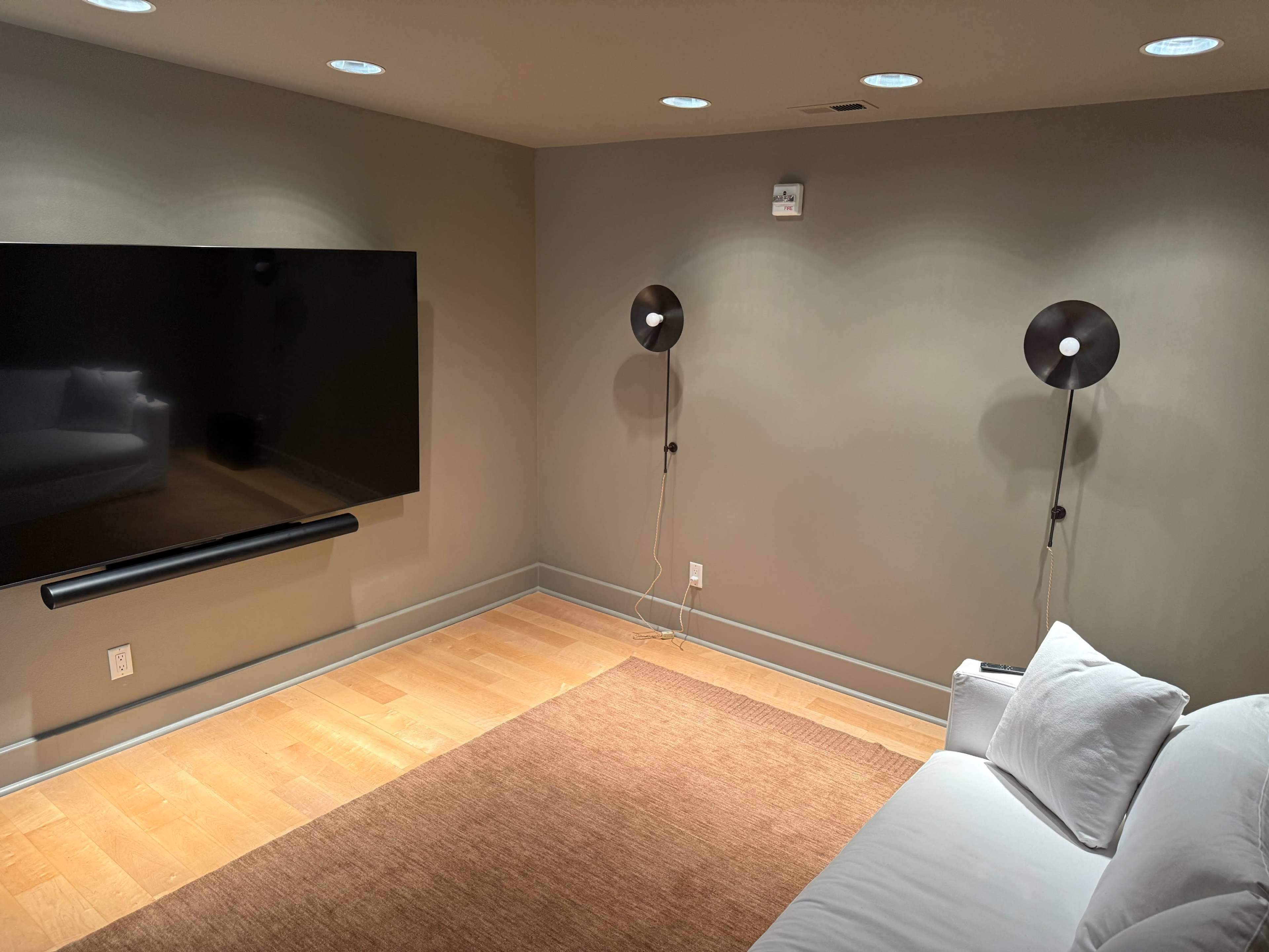The image shows a modern, minimalistic living room featuring a large flat-screen television mounted on the wall, two sconces on either side, and a white sofa facing the TV over a beige area rug.
