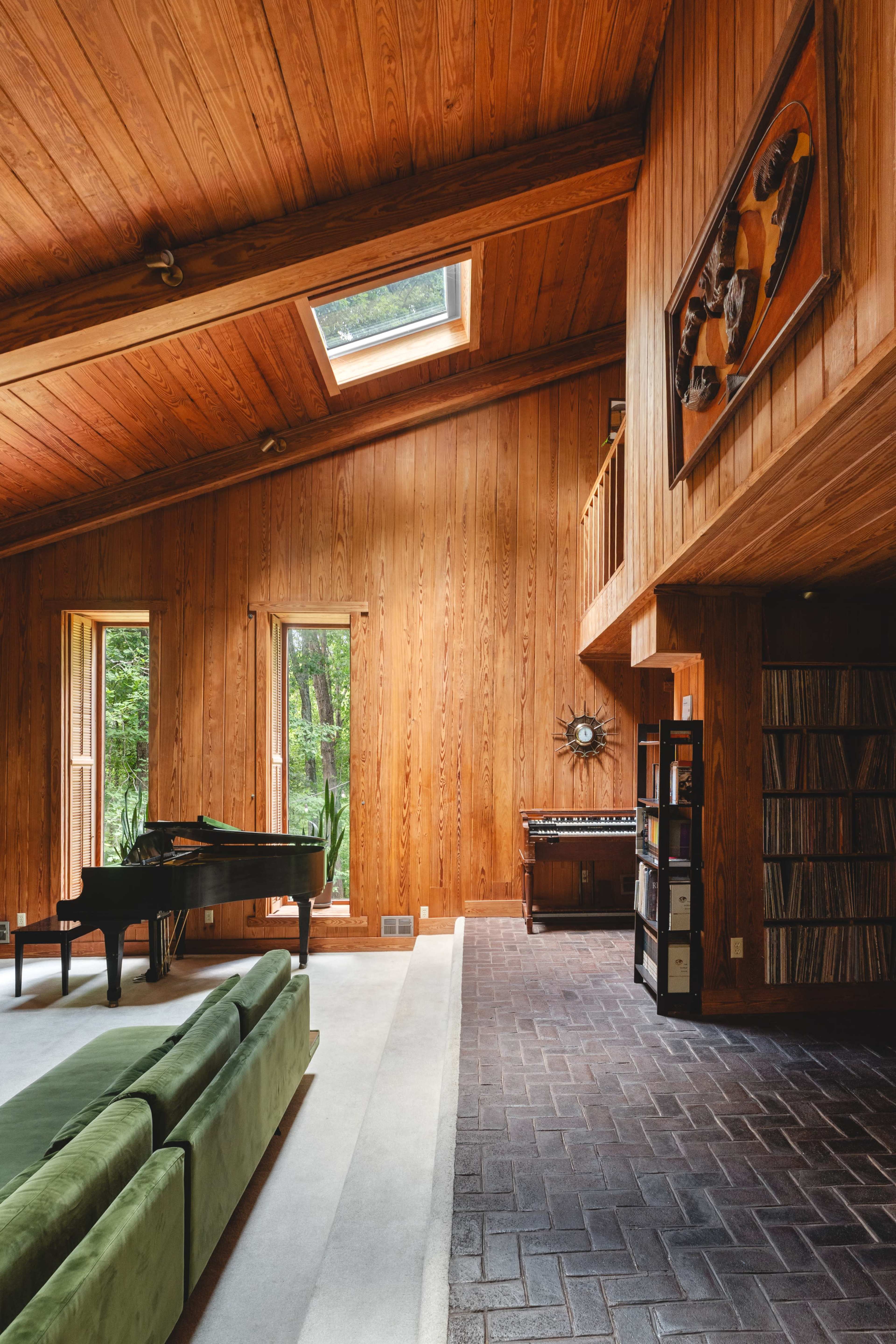 The image shows a wooden-paneled living room featuring a grand piano, a green sofa, and large windows that overlook a natural setting.