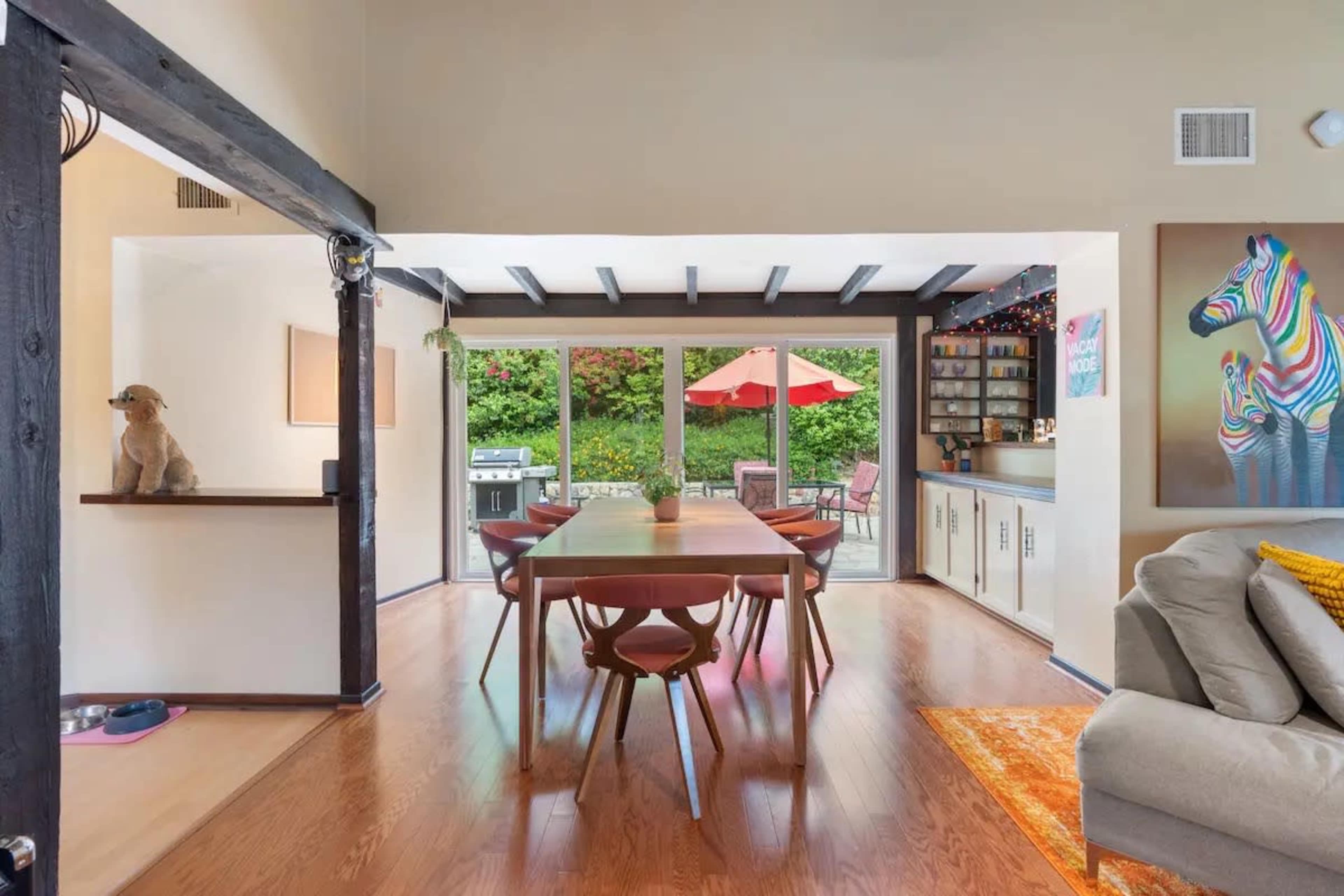 The image shows a modern dining area with a long wooden table surrounded by chairs, leading to a spacious outdoor patio visible through large glass doors.