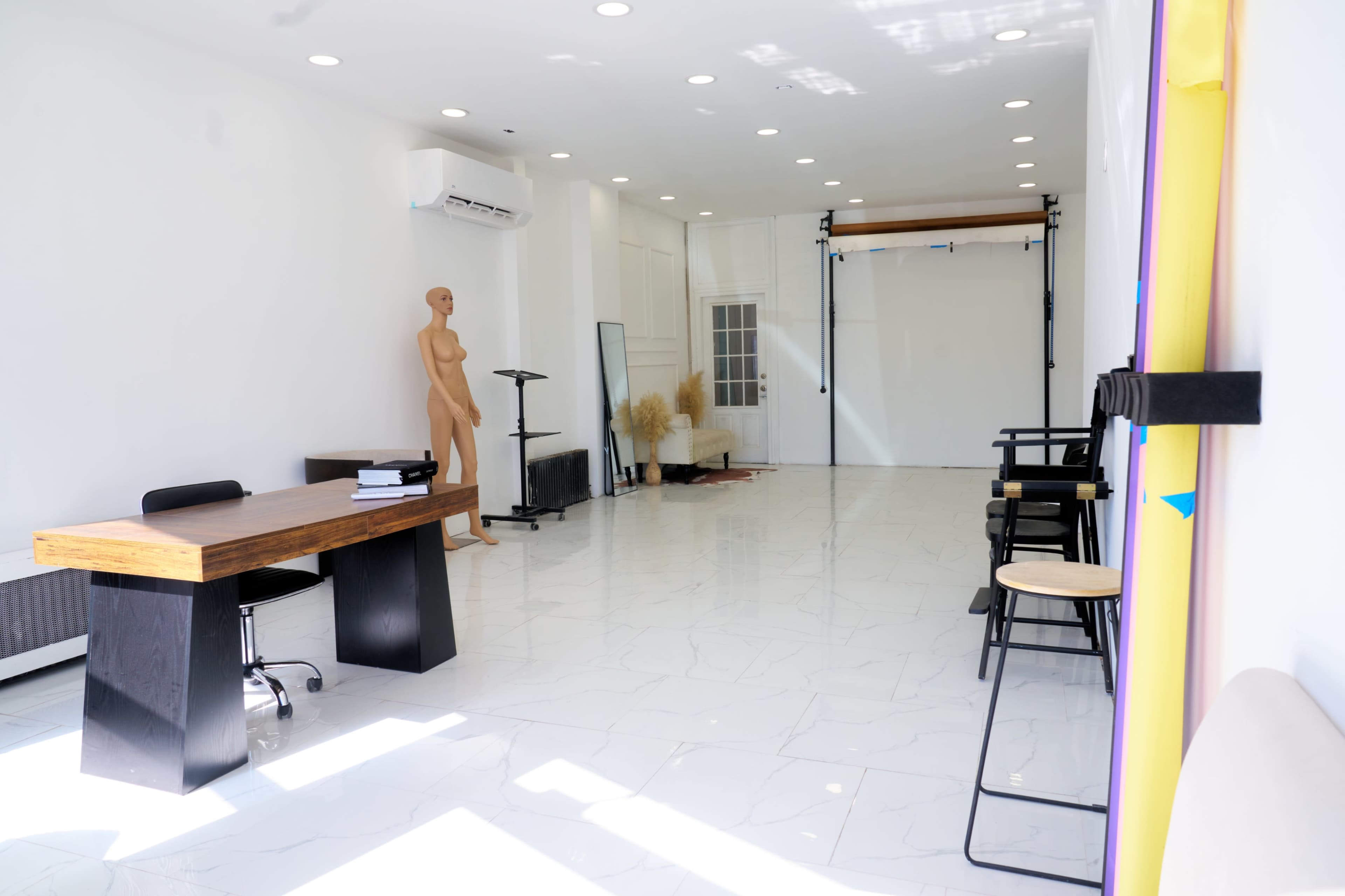 The image shows a spacious, well-lit studio featuring a wooden desk, a mannequin, a mirror, and minimalist furniture against a white wall.