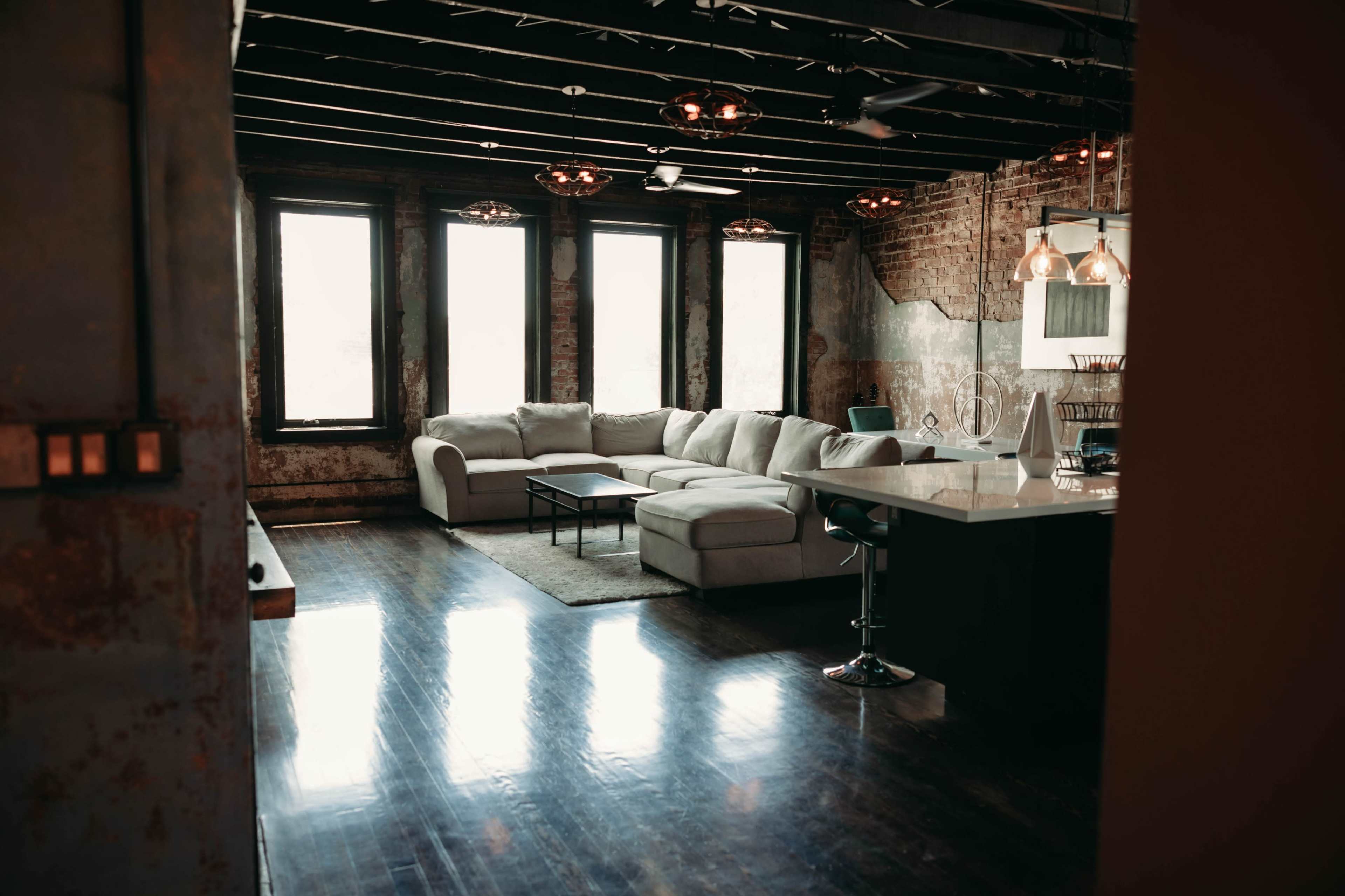The image shows a modern living space with a sectional sofa, a coffee table, and large windows, featuring exposed brick walls and a kitchen area with bar seating.
