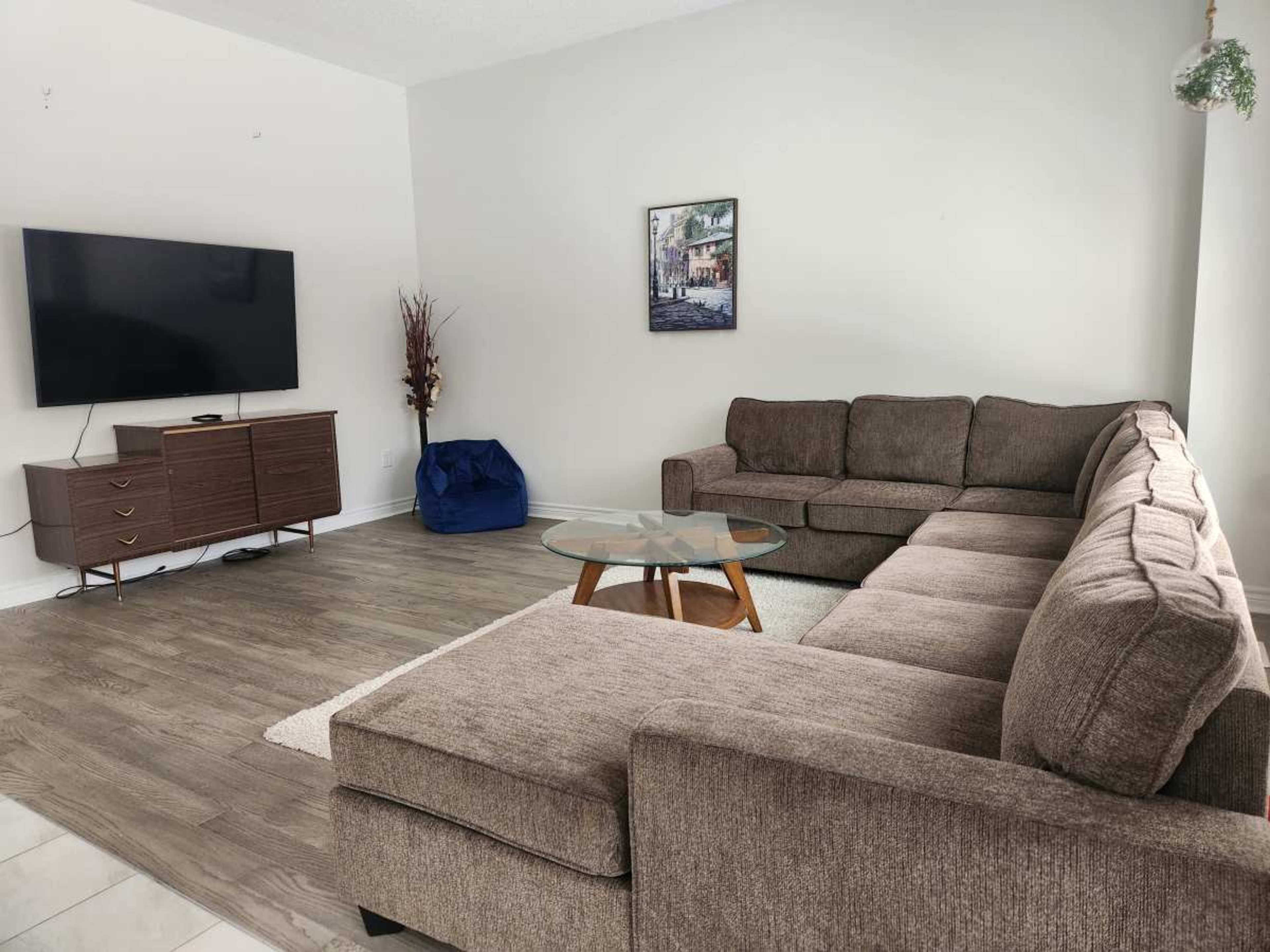 The image shows a modern living room featuring a sectional sofa, a glass coffee table, a television mounted on the wall, and a wooden cabinet.