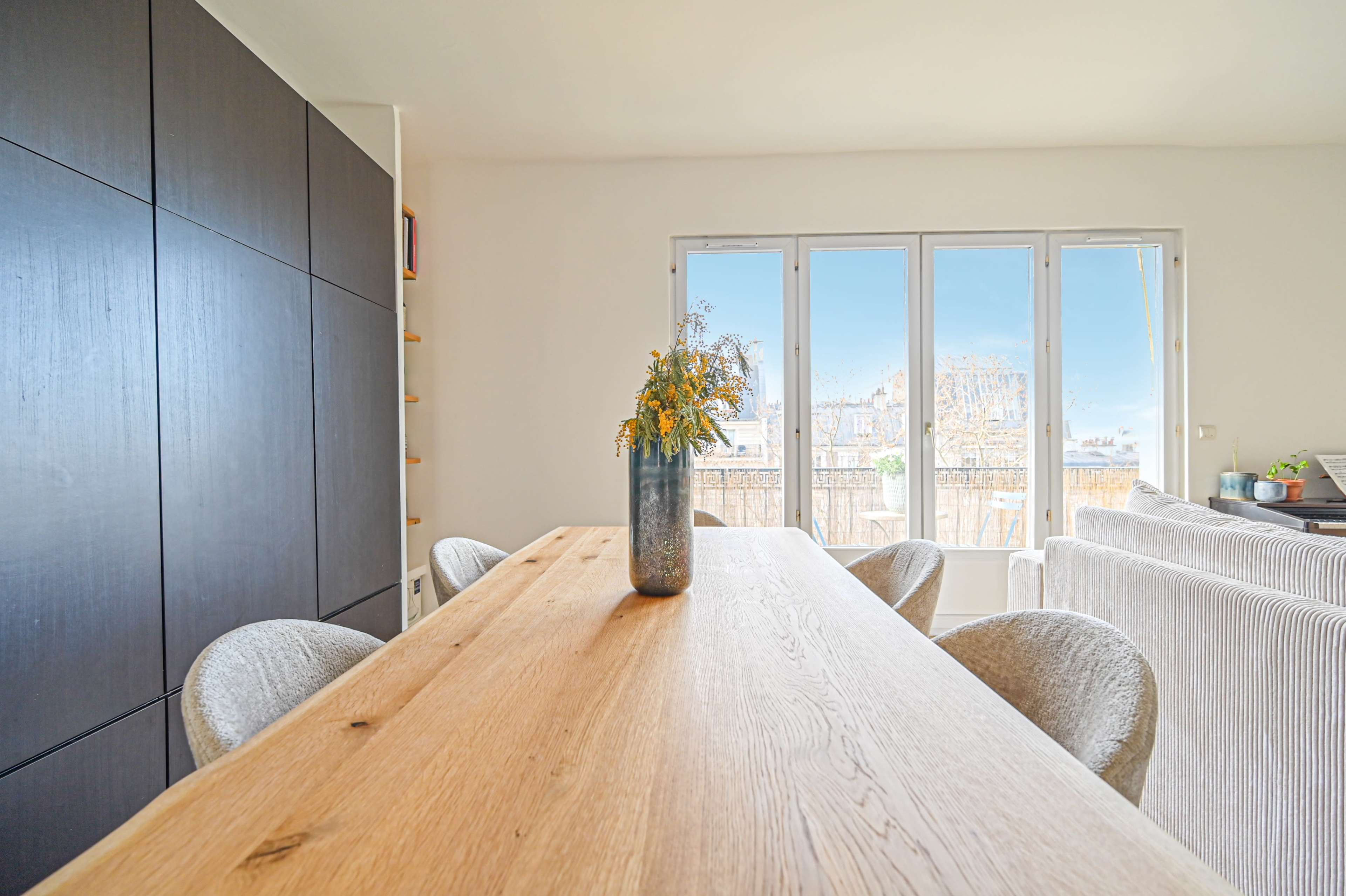 A dining table with wooden surface and six chairs is set against a backdrop of large windows overlooking a cityscape, with a vase of flowers at the center.