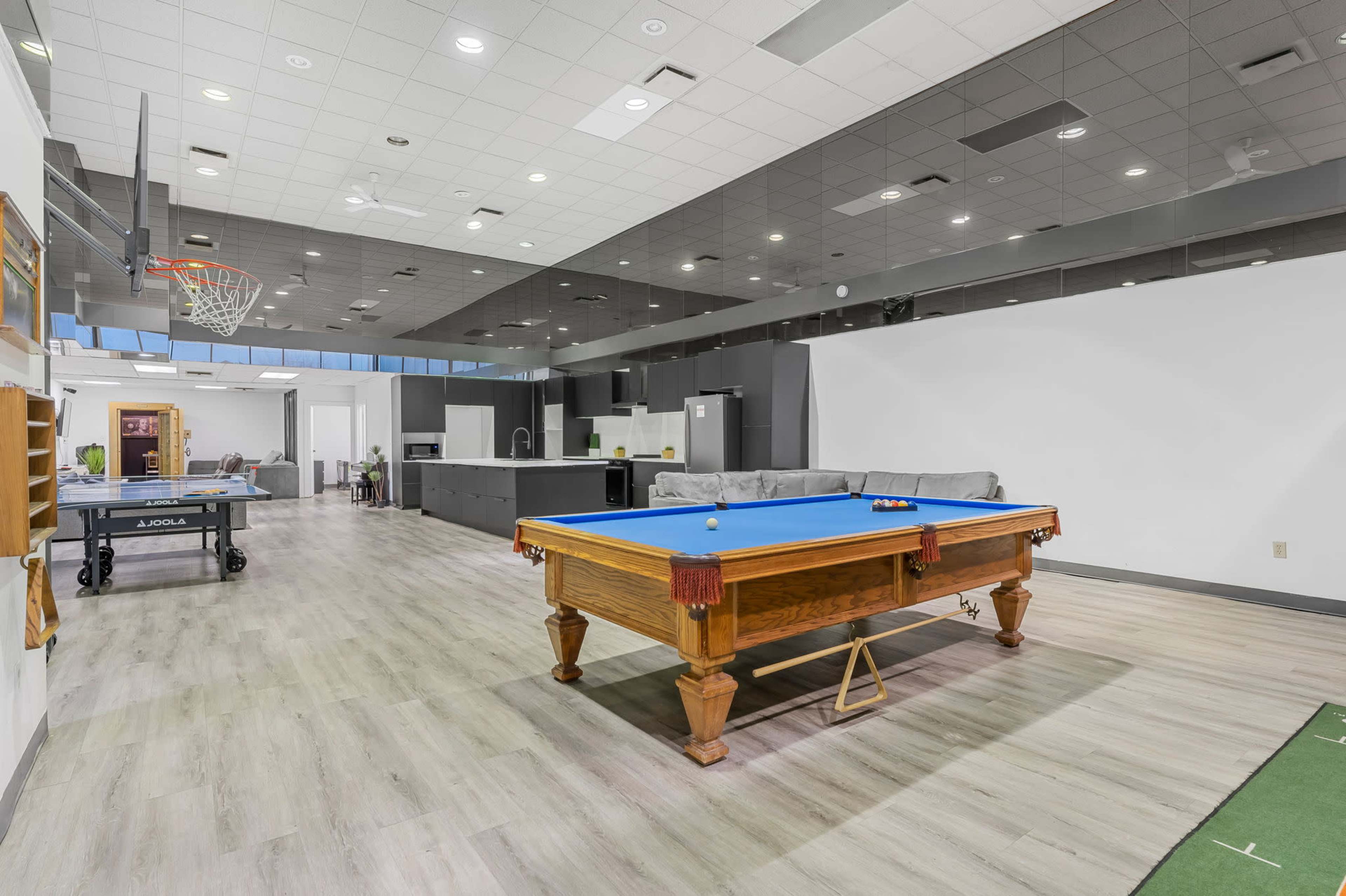 The image shows a spacious game room featuring a blue pool table, a basketball hoop, and a modern kitchen area in the background.