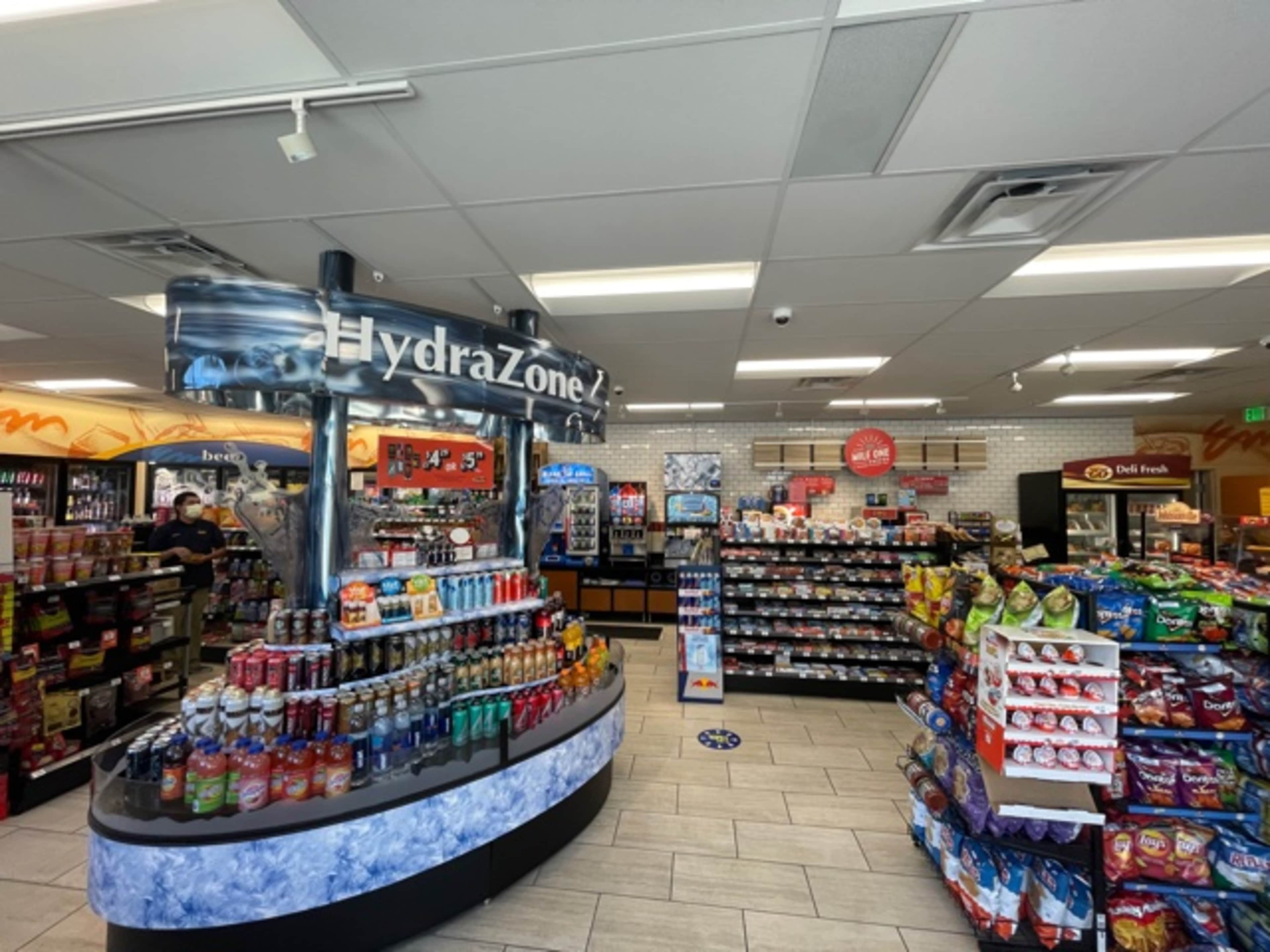 The image shows a convenience store interior with a central display of beverages labeled "HydraZone" and shelves stocked with snacks and pre-packaged food items.