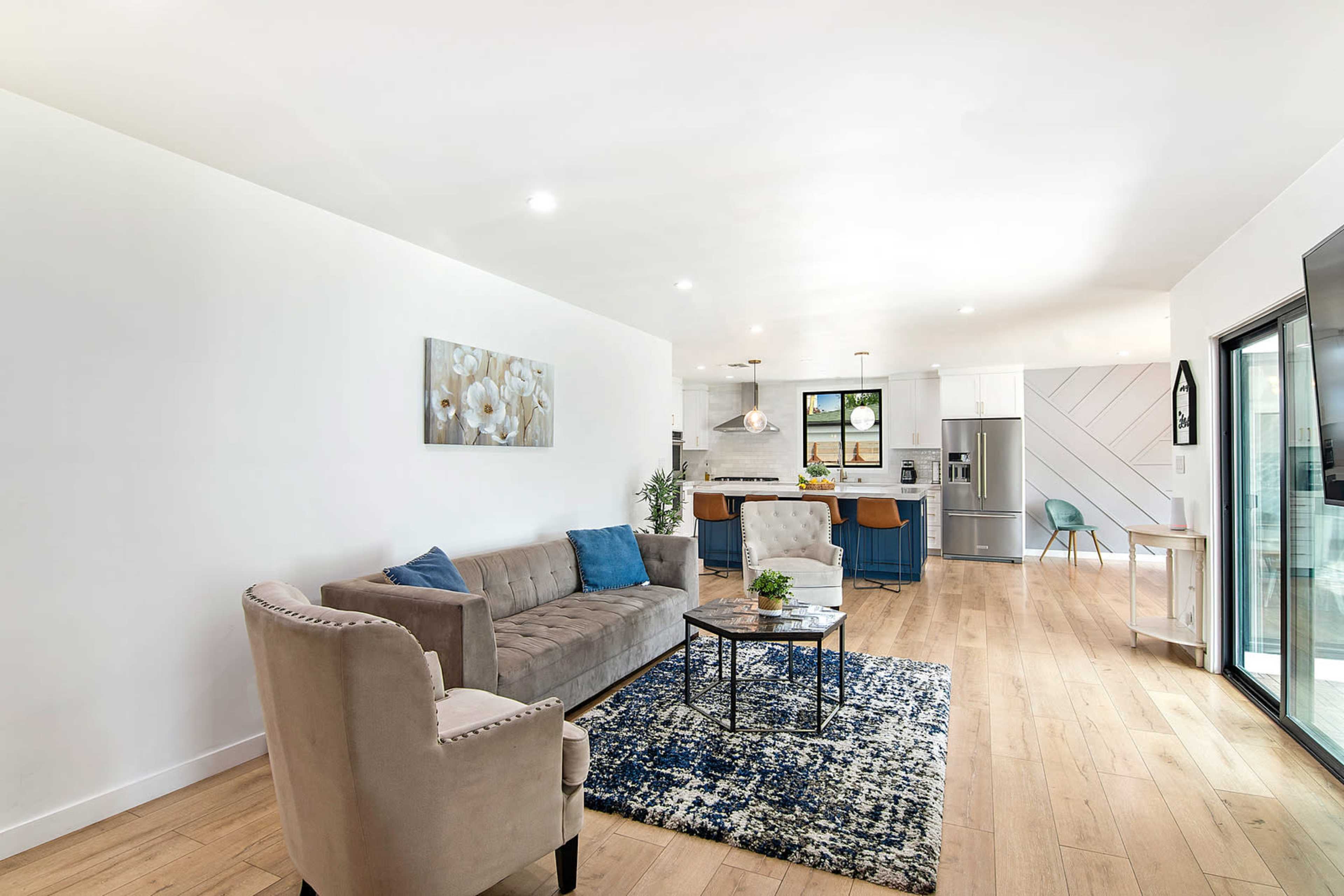 A modern living room features a gray sofa and an armchair, with a coffee table on a blue patterned rug, leading to a kitchen area with blue cabinets and stainless steel appliances.