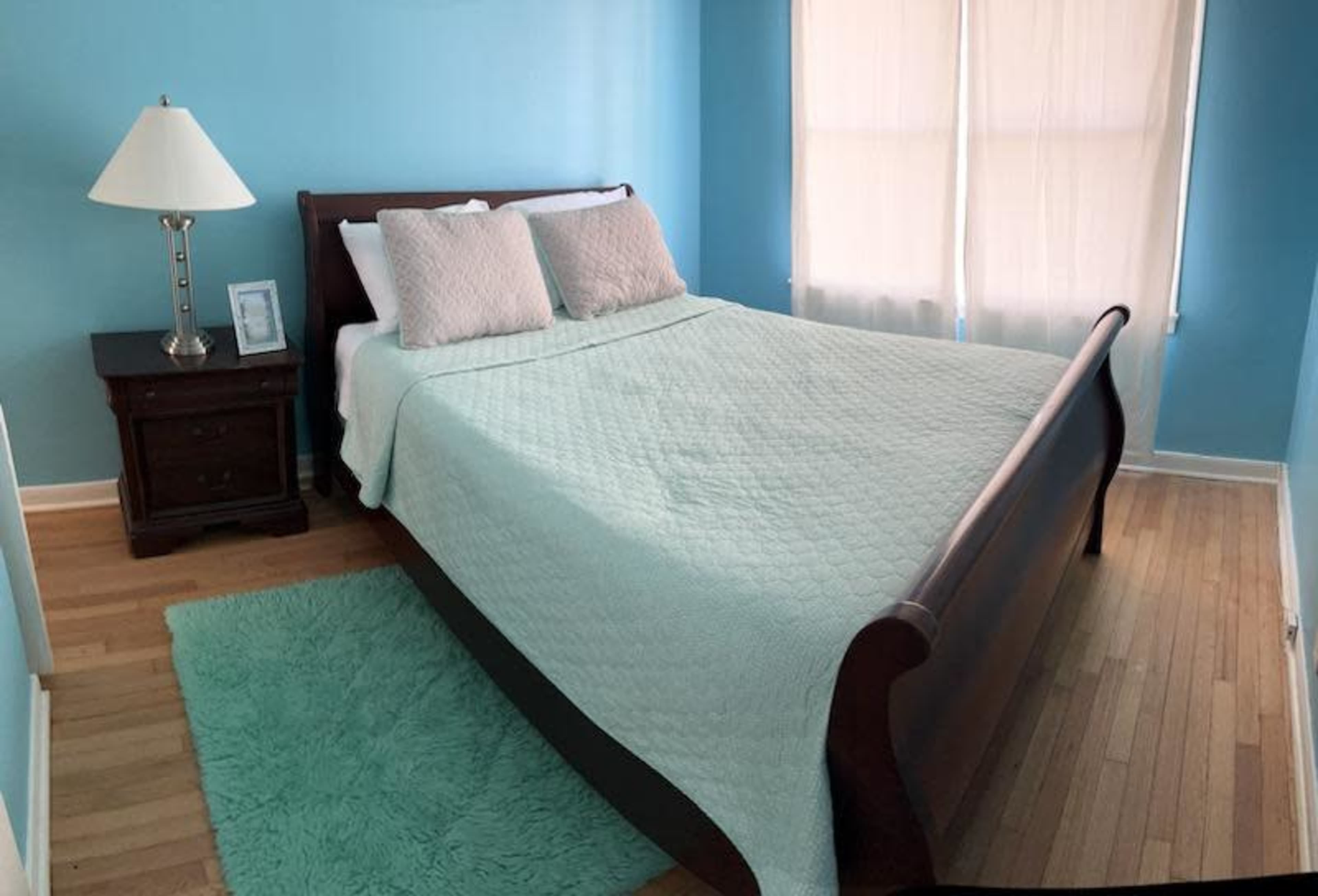 The image shows a bedroom with a wooden sleigh bed covered in a light quilt, a nightstand with a lamp, and large windows with sheer curtains on a hardwood floor.