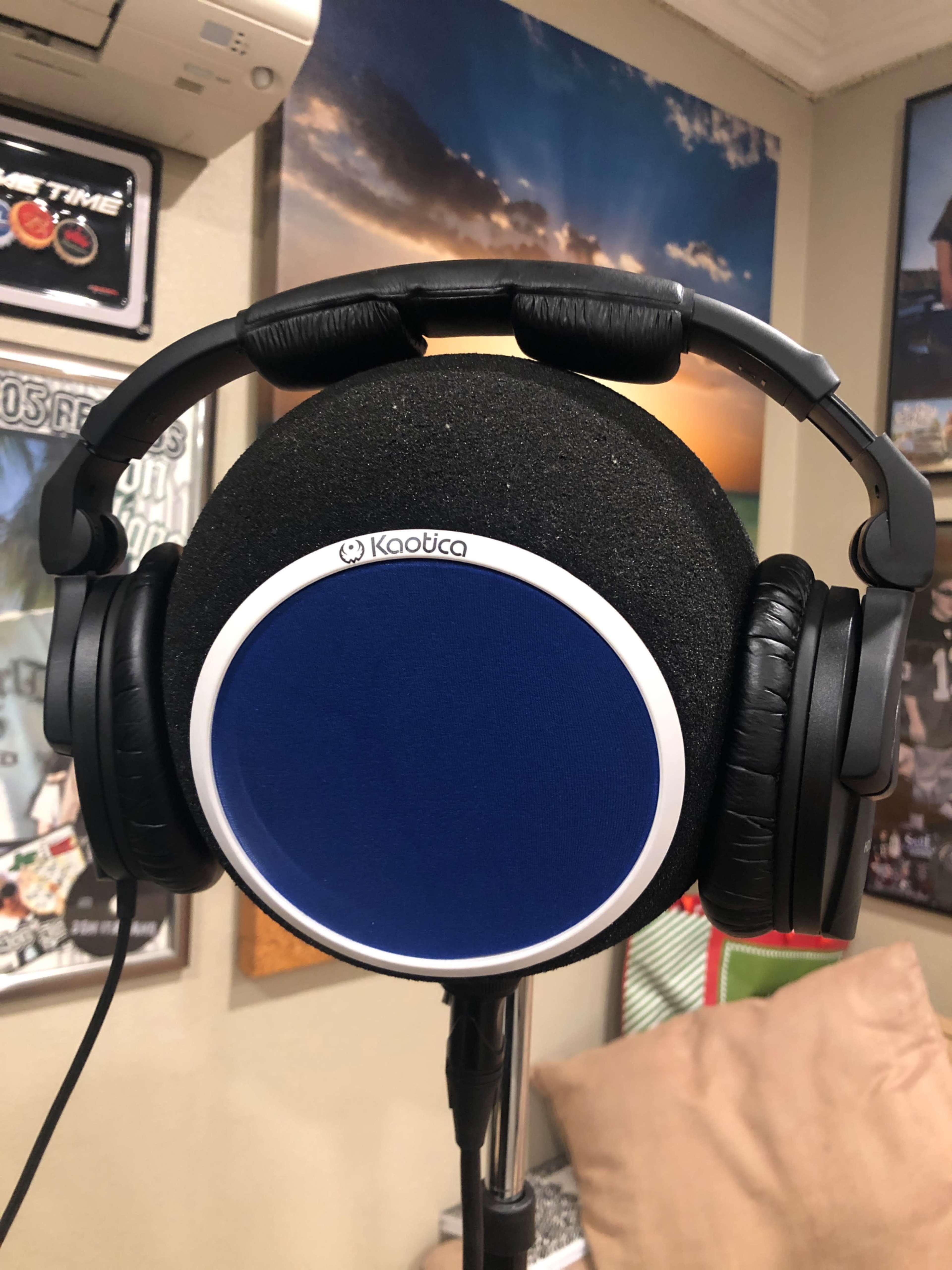A pair of black headphones rests on a blue pop filter mounted on a microphone stand against a backdrop of posters and cushions.
