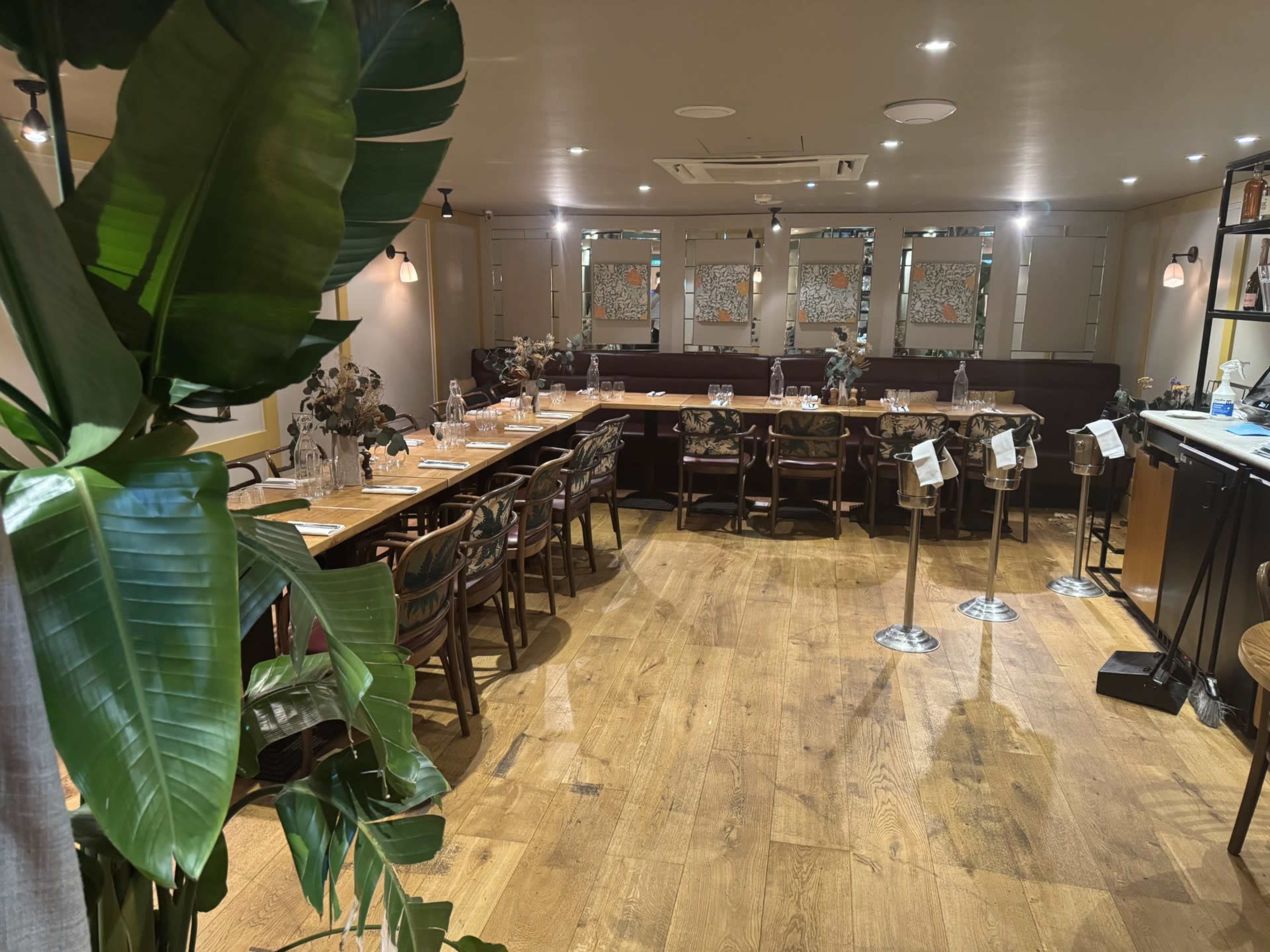 The image shows a restaurant interior with wooden floors, a long dining table set with glassware, and a bar area in the background.